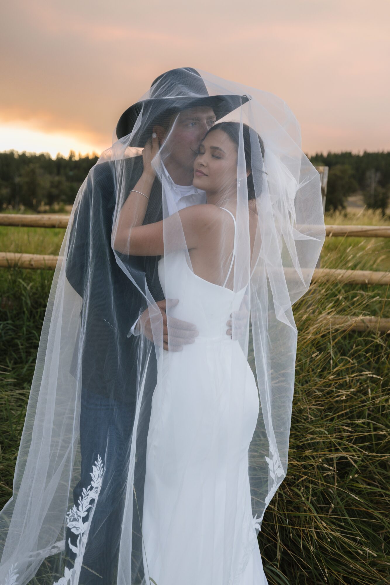 A bride and groom embrace outdoors, with the bride in a white dress and veil, and the groom in dark clothing, in a field.