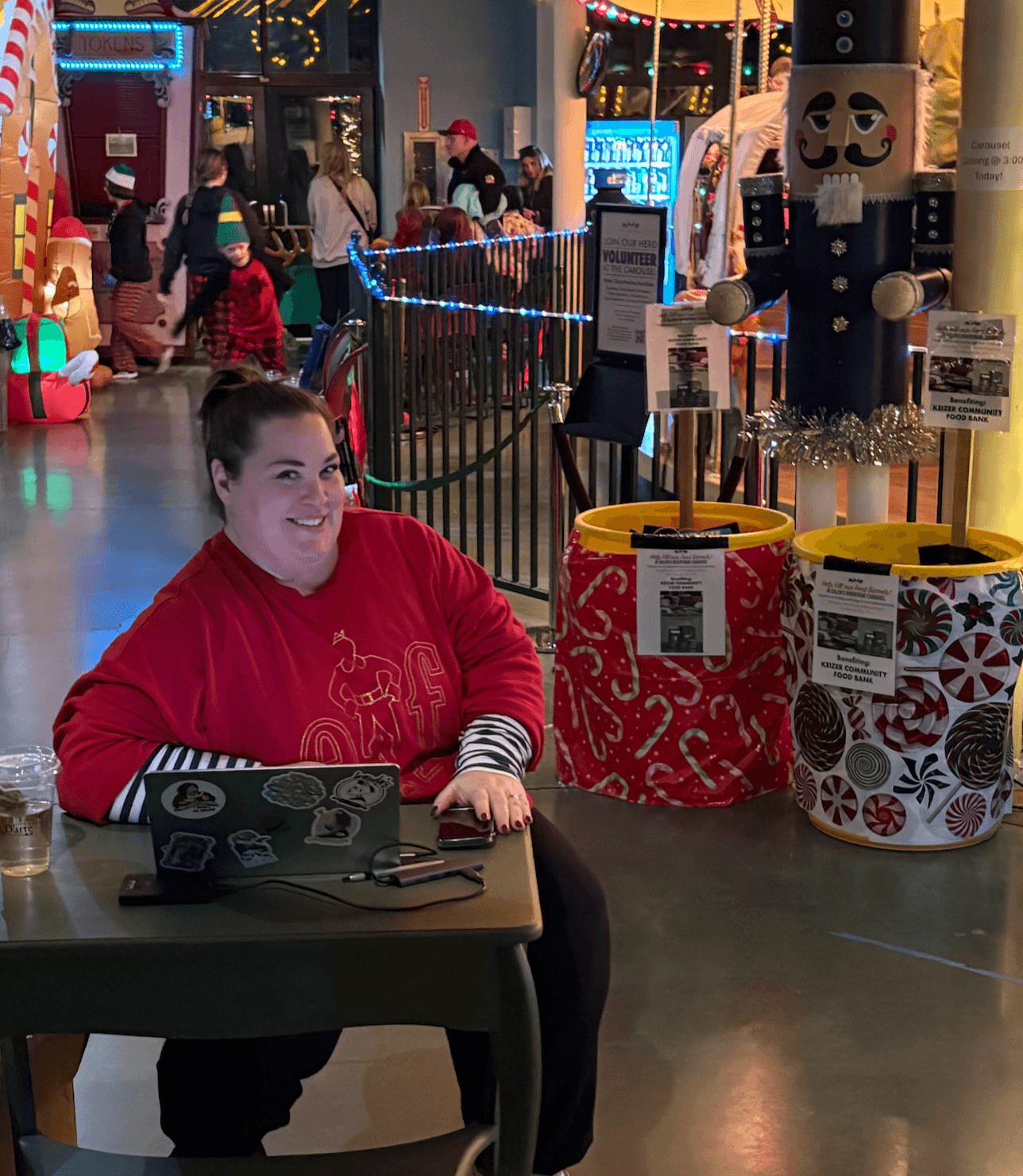 Woman sitting at a table in an indoor amusement area, smiling, with festive decorations and colorful lights in the background.