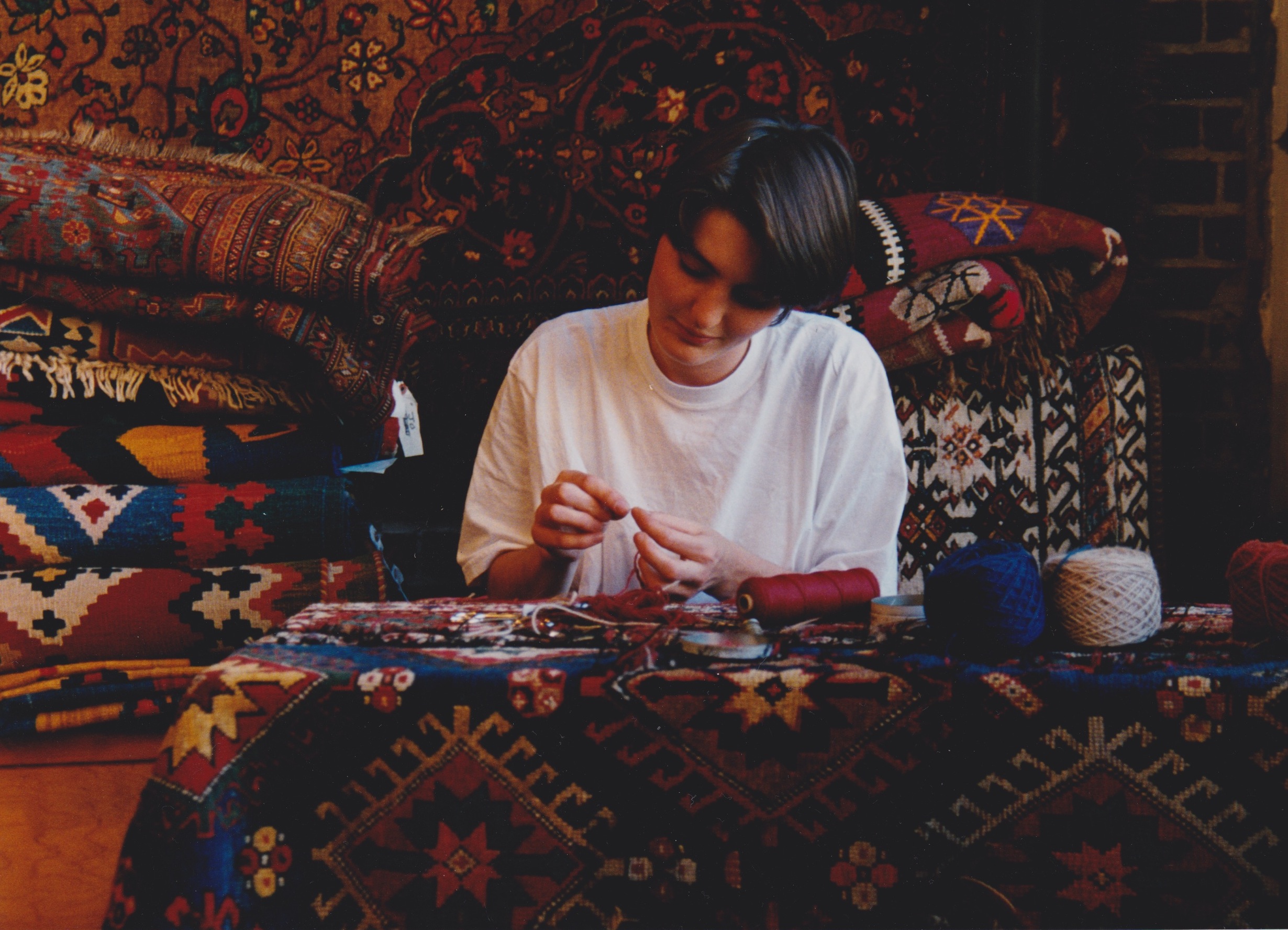 Person with short dark hair sitting at a table, working on a craft project, surrounded by colorful textiles and cushions