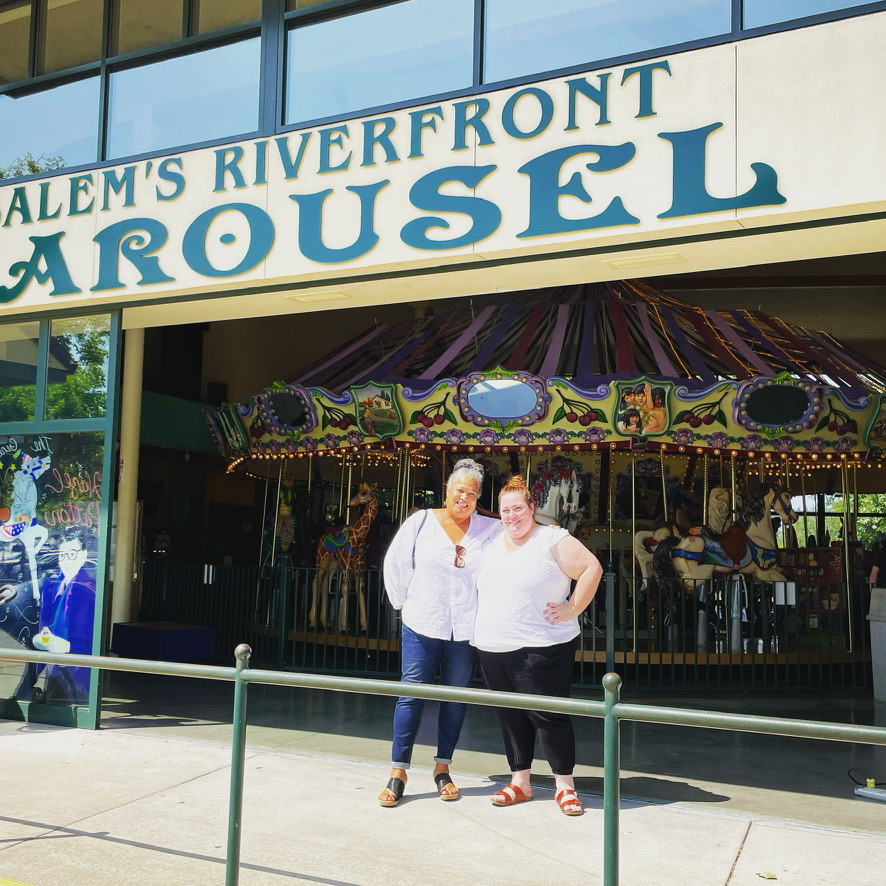 Two women stand in front of a carousel at Salem's Riverfront, smiling. The carousel is decorated with colorful lights and patterns.