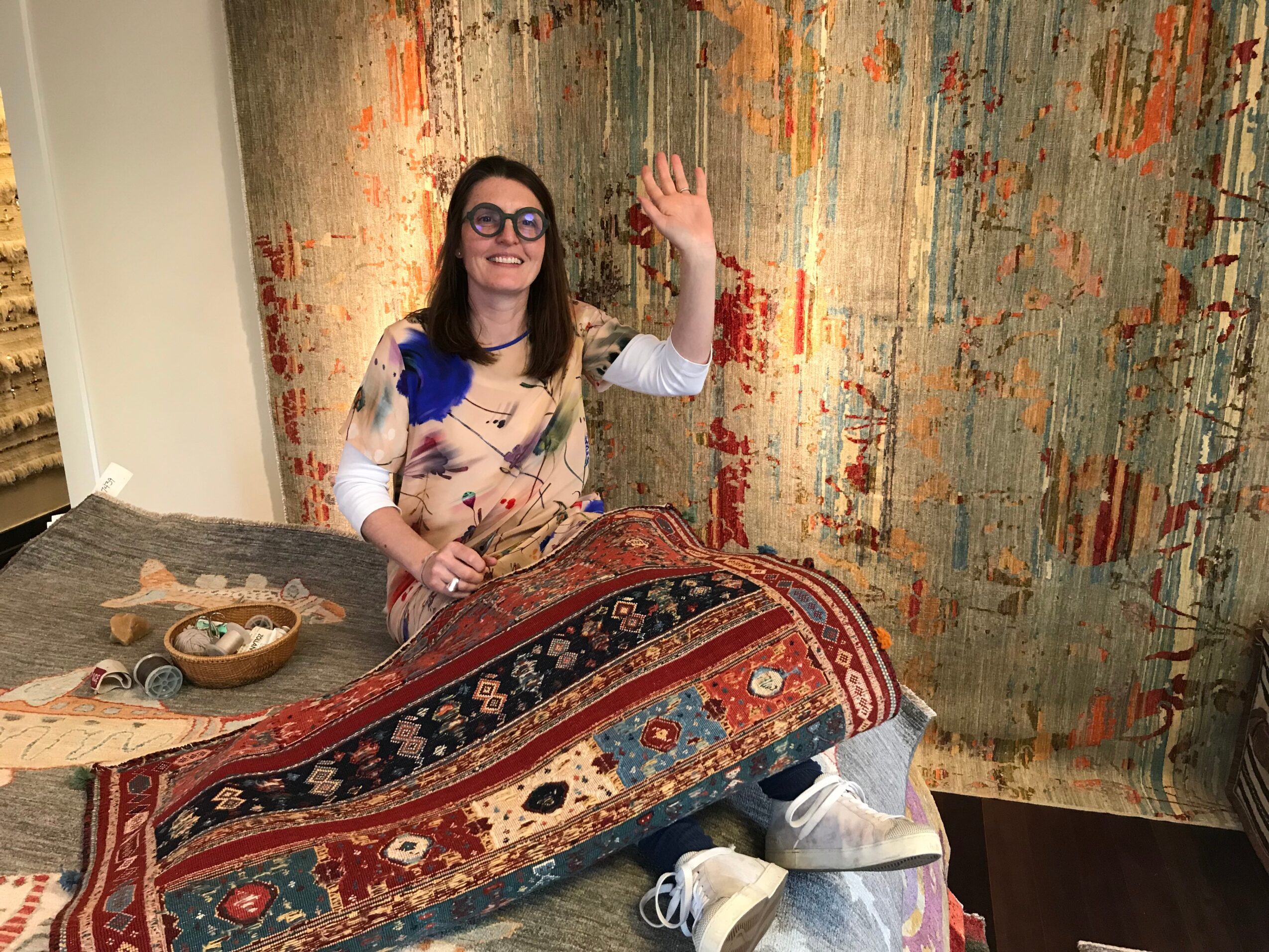 Woman with glasses and long hair, smiling and waving, standing behind a table with a colorful rug, in front of an abstract wall art.