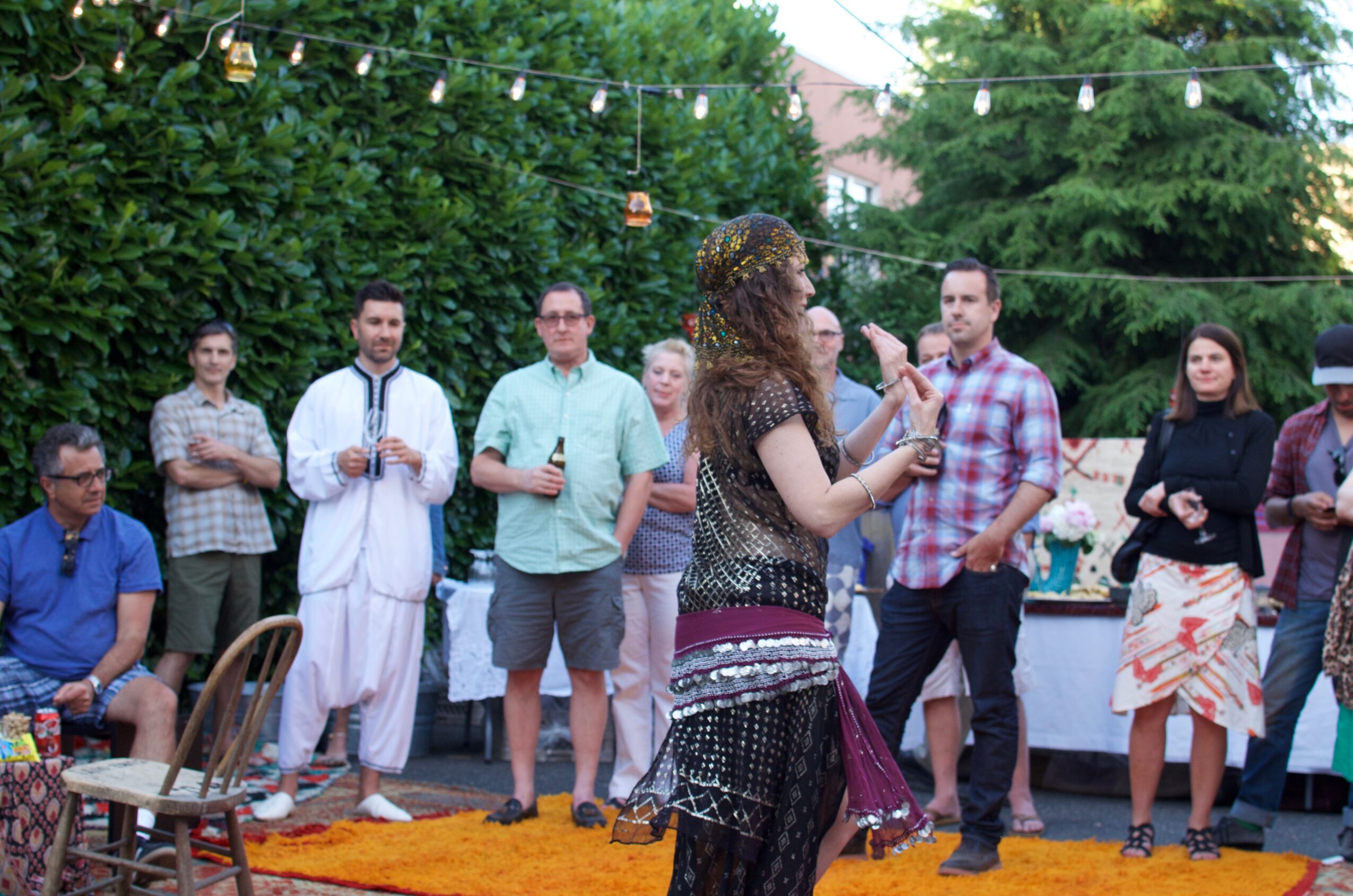 Group of people outdoors, some standing and some sitting, with a woman speaking into a microphone, trees, and string lights in background.