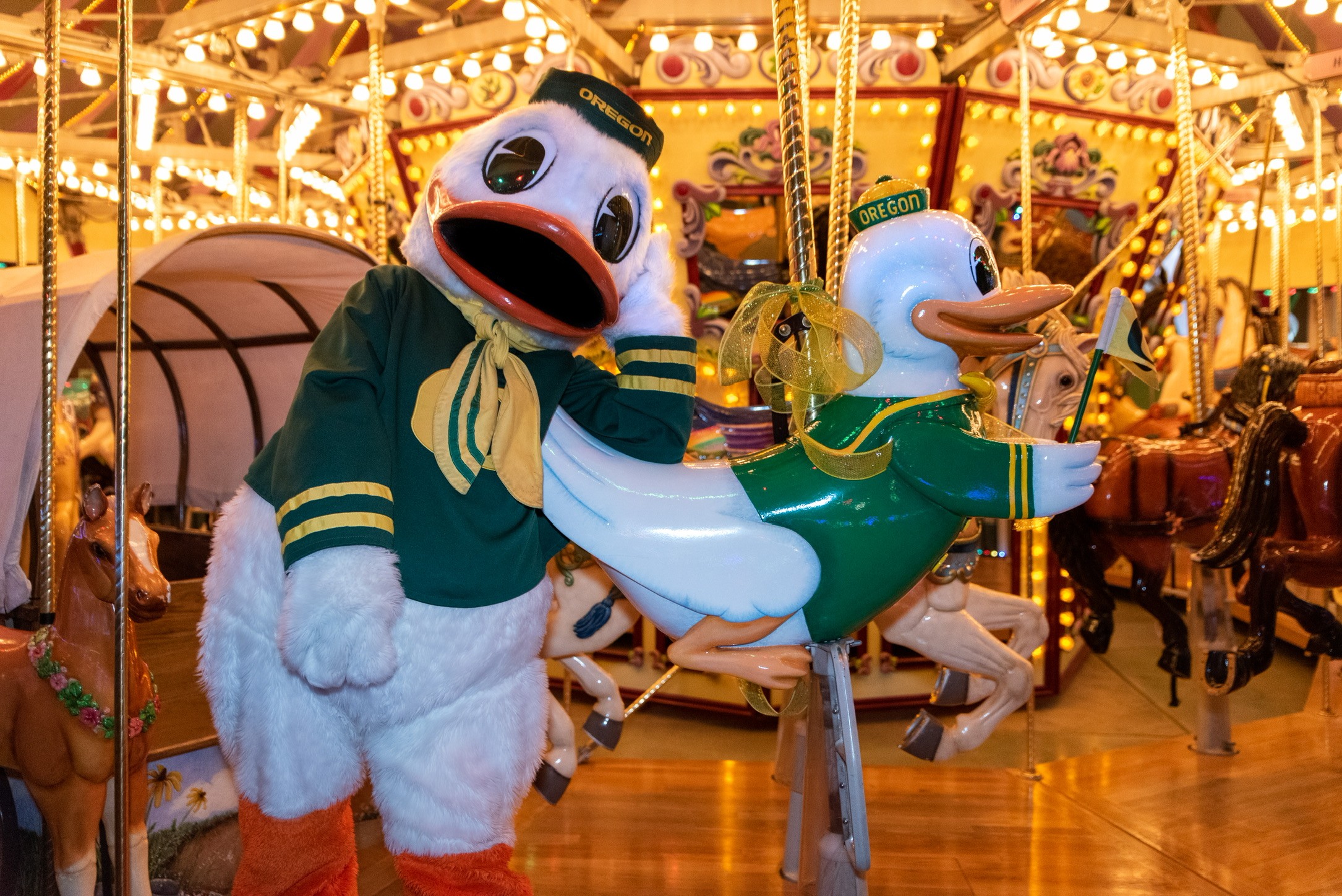 Person in a duck mascot costume standing next to a carousel duck ride, amusement park background with lights.