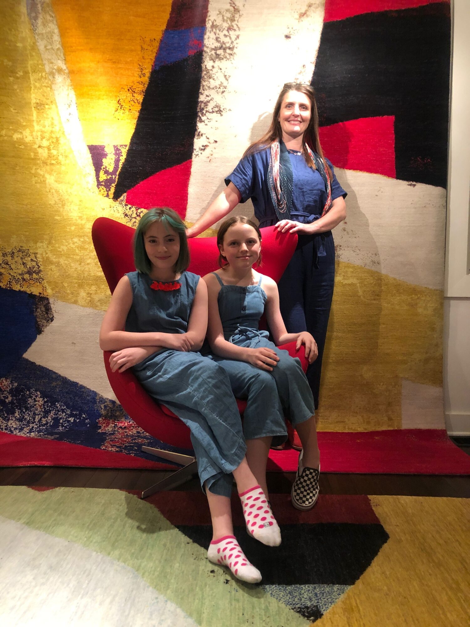 Three women sitting on a red chair in a room with colorful abstract carpet, two women sitting on the chair, one standing behind.