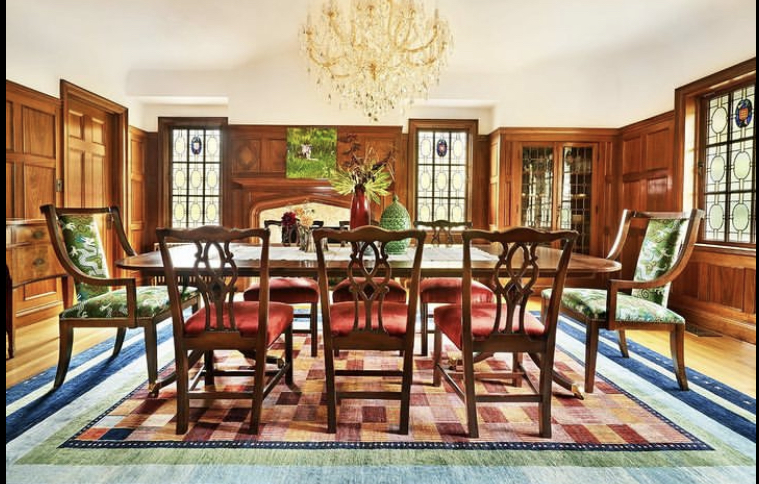 Elegant dining room with wooden furniture, large windows, a chandelier, and a colorful rug under the table.