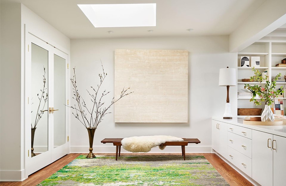 Bright room with a bench, large wall art, white cabinet, and decorative plants, illuminated by a skylight.