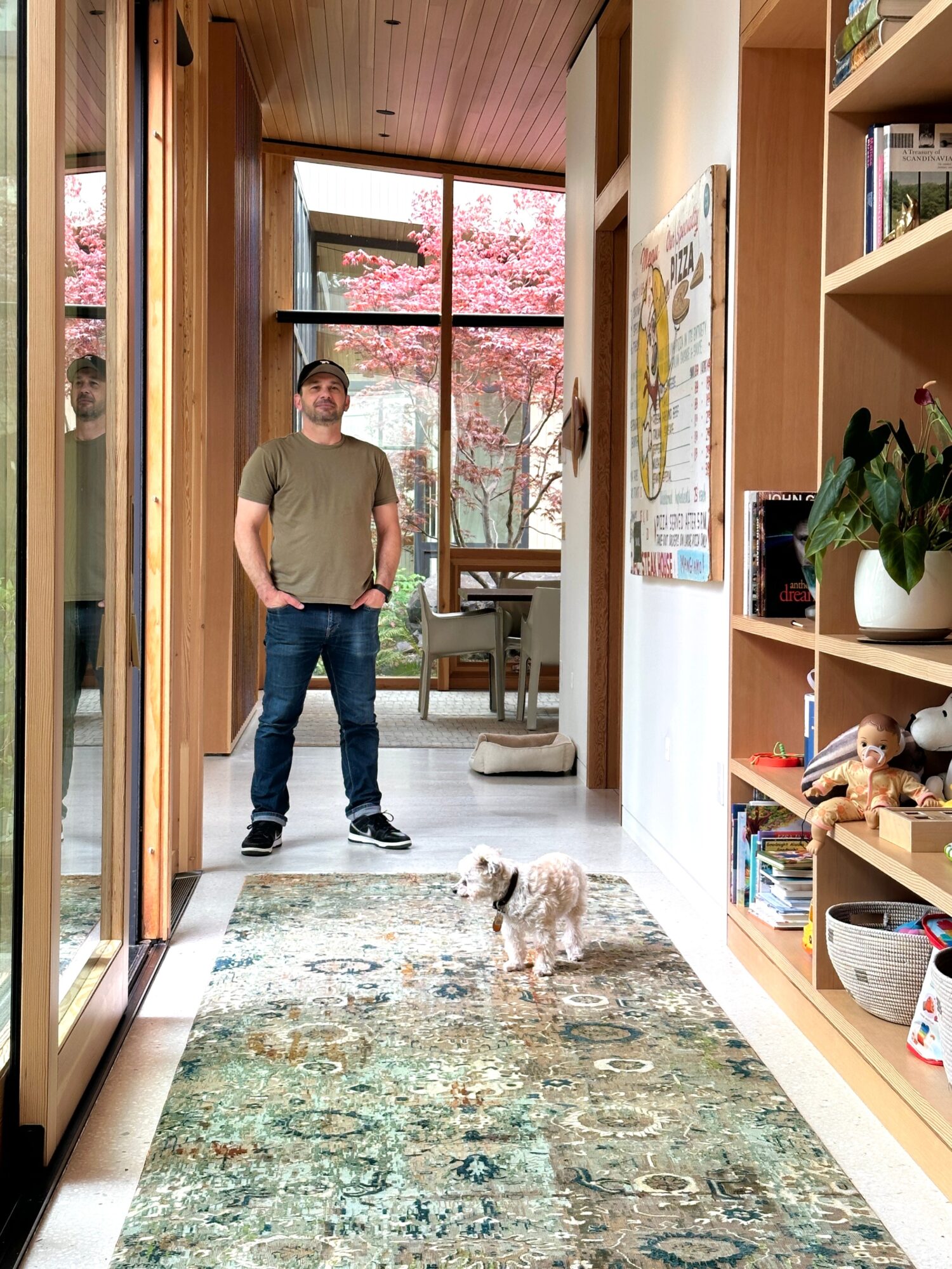 Man standing in a hallway with a small dog on a patterned rug, bookshelves on the right, and a window with pink trees outside.