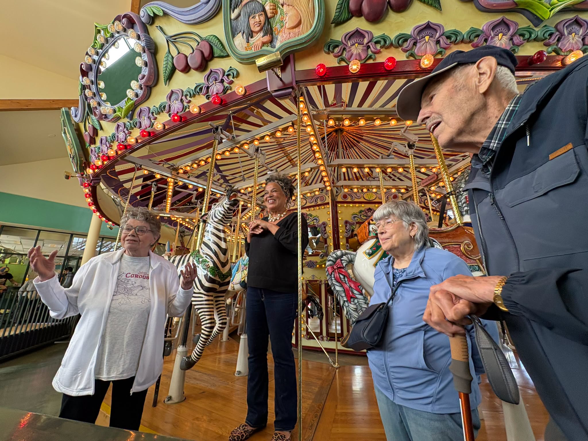 Group of four older adults near a carousel with a zebra, some holding drinks, in an indoor amusement setting.