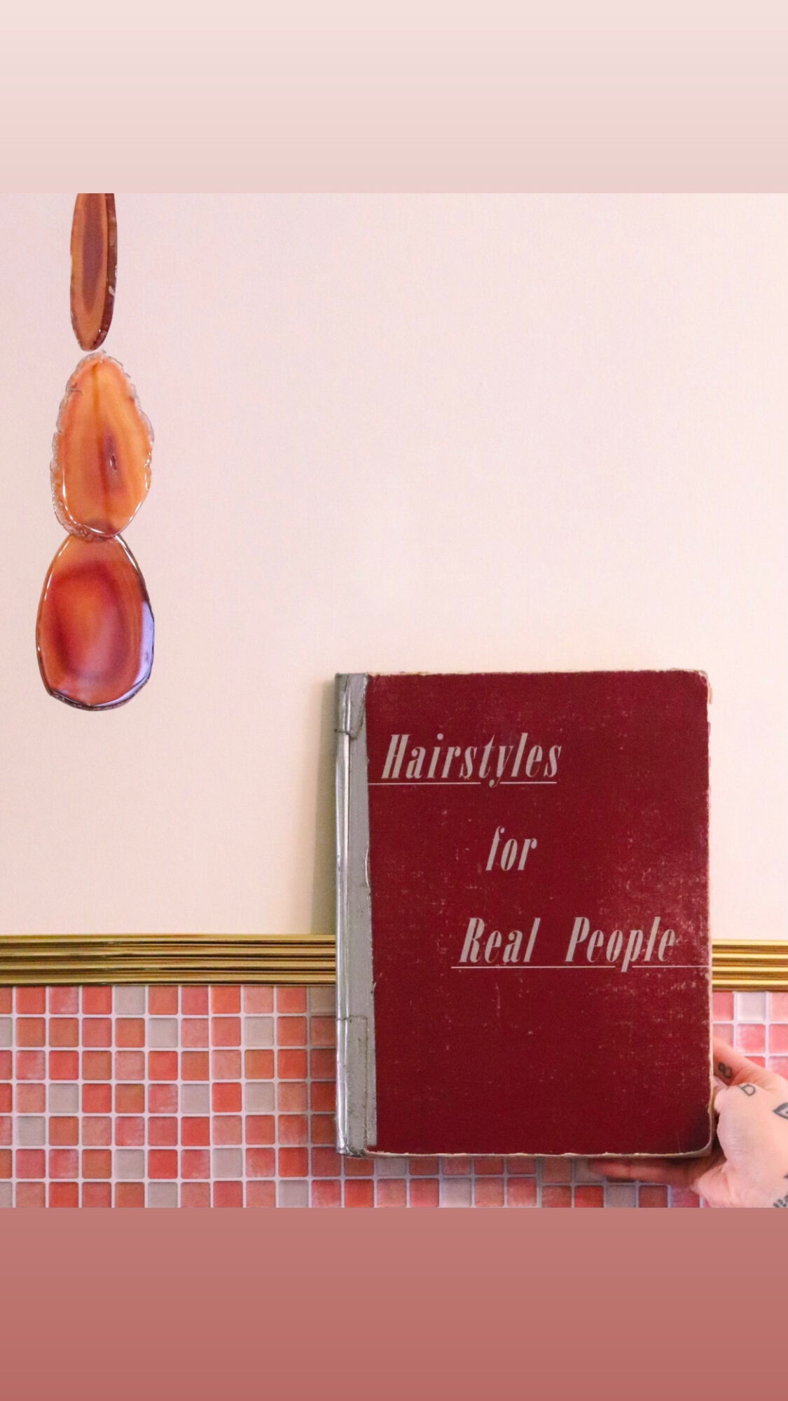 A book titled 'Hairstyles for Real People' leaning against a wall with a pink and red tiled pattern below.