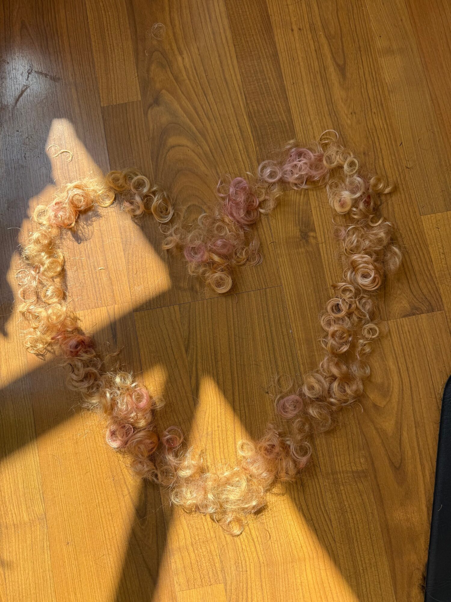 Heart-shaped arrangement of curly hair extensions on wooden floor, with sunlight and shadows, and a partial view of a black object.