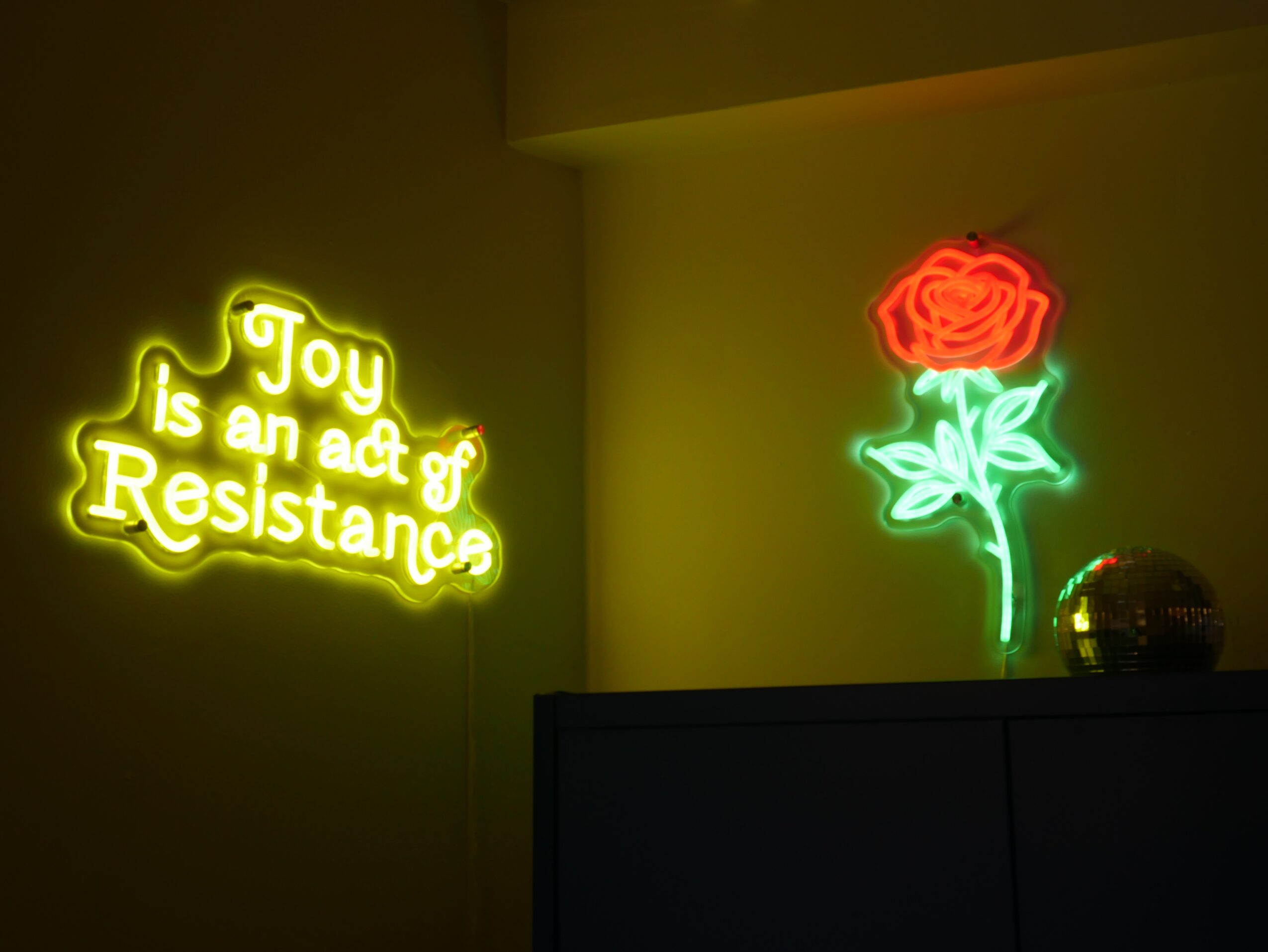 Neon sign reads 'Joy is an act of Resistance' in yellow, and a neon red rose with green leaves on a wall.