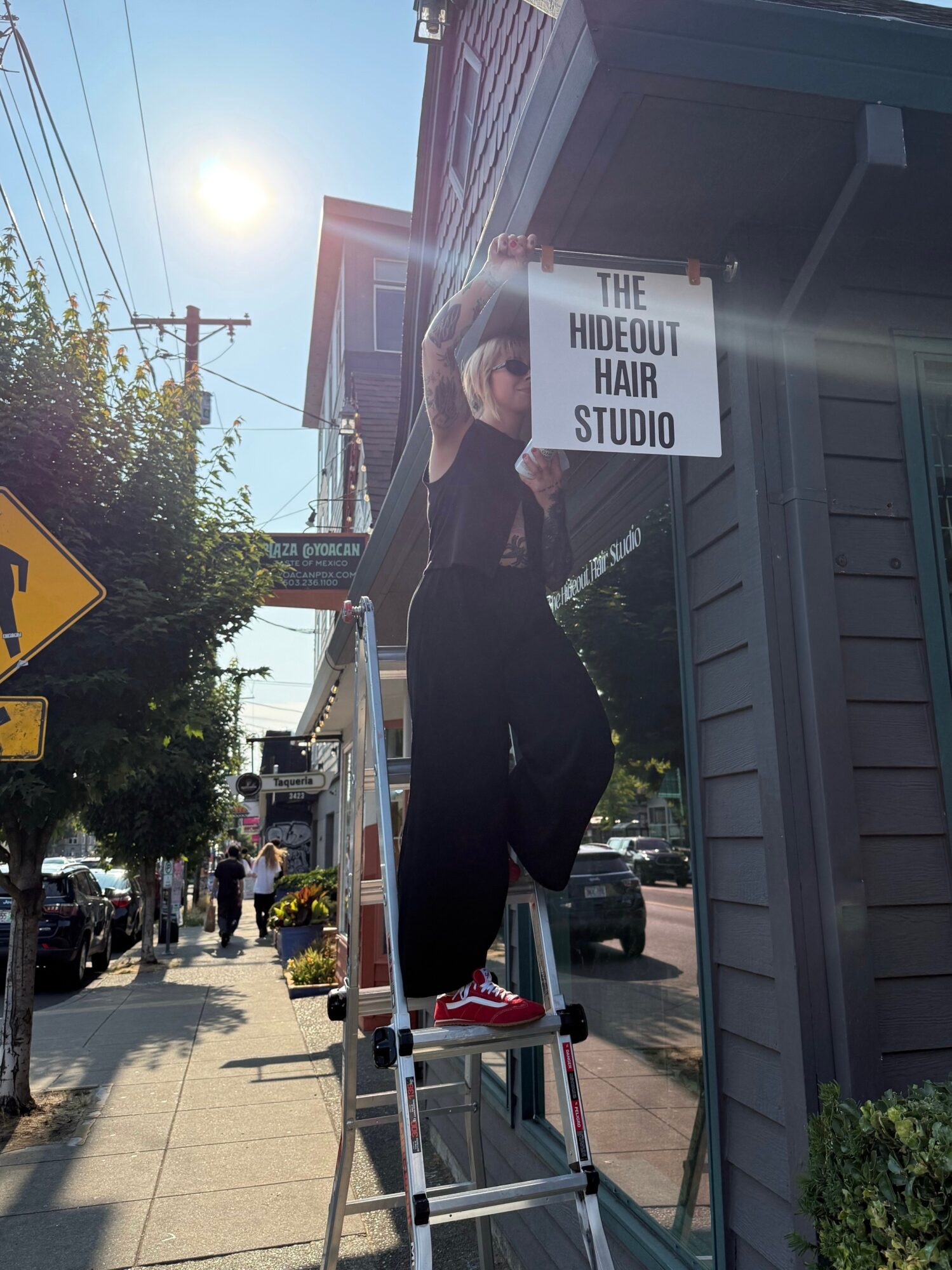 Person on ladder hanging sign for The Hideout Hair Studio on building exterior, sunny day, sidewalk scene.