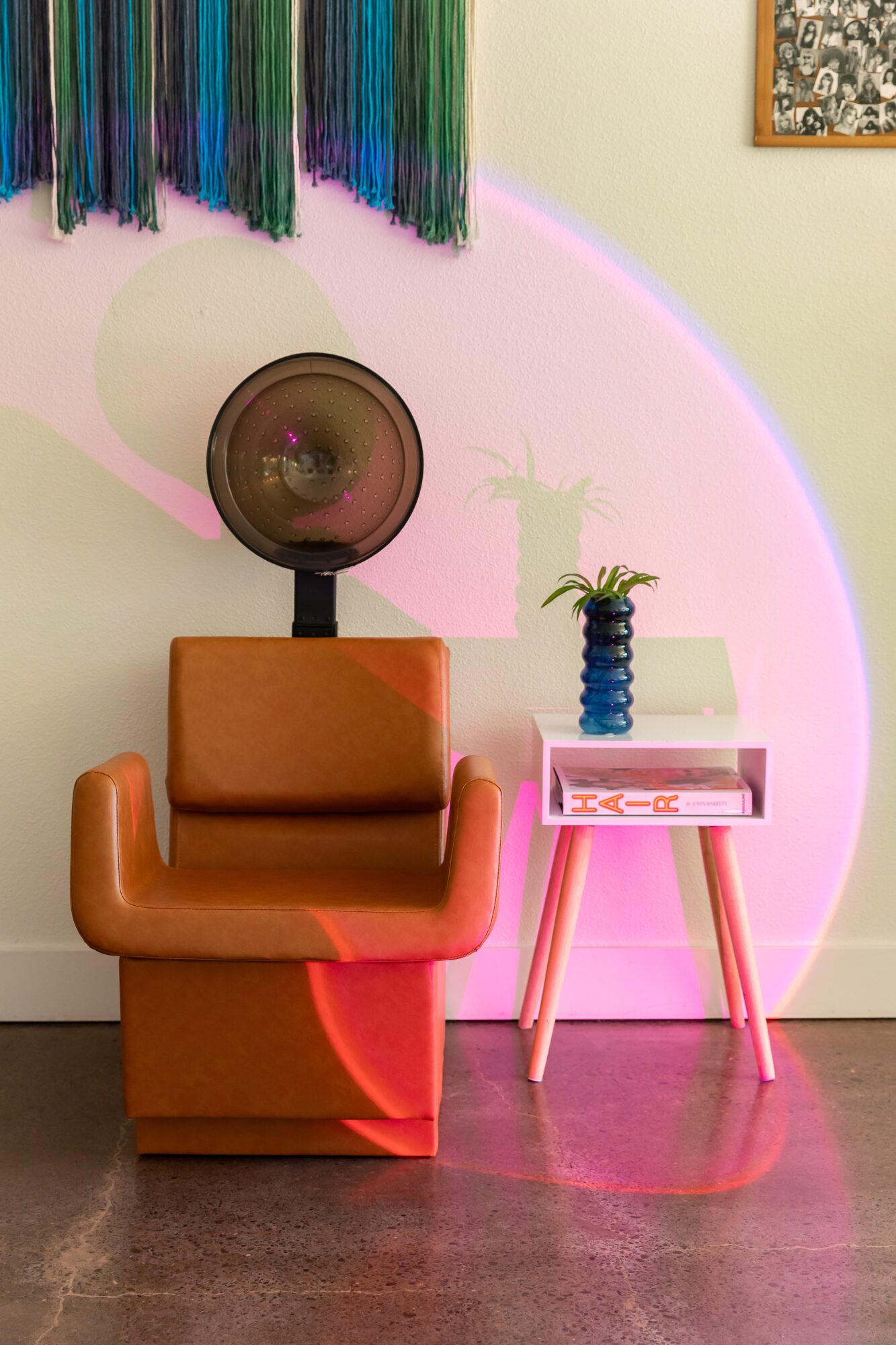 Room corner with colorful wall hanging, vintage fan, orange armchair, pink side table, and potted plant, illuminated by pink light.