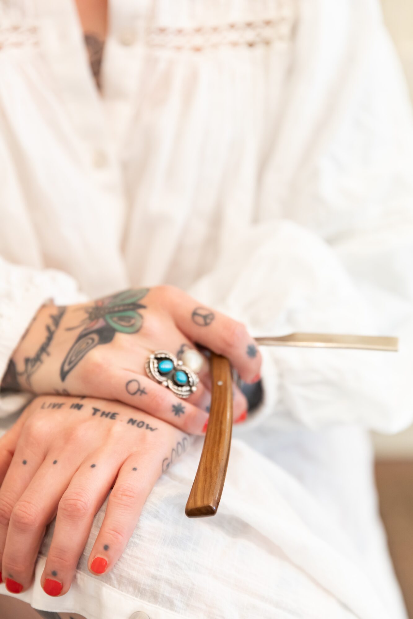 Close-up of hands with tattoos, rings, and red nail polish, holding a wooden-handled tool against a white fabric background.
