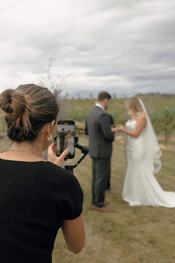 Person taking a photo of a wedding couple outdoors on cloudy day, woman with brown hair in bun, holding phone, couple in formal attire, bride in white dress, groom in dark suit, grassy field background.