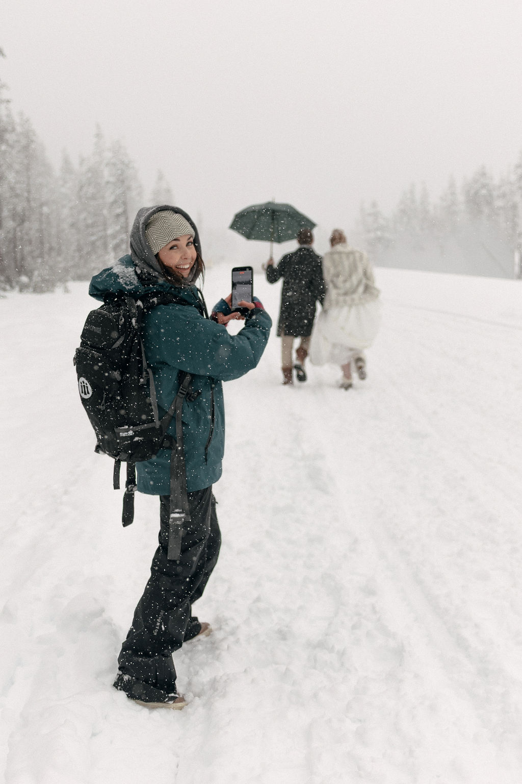 Person in winter clothing taking a photo with a phone in snowy landscape, others walking with umbrella in background.