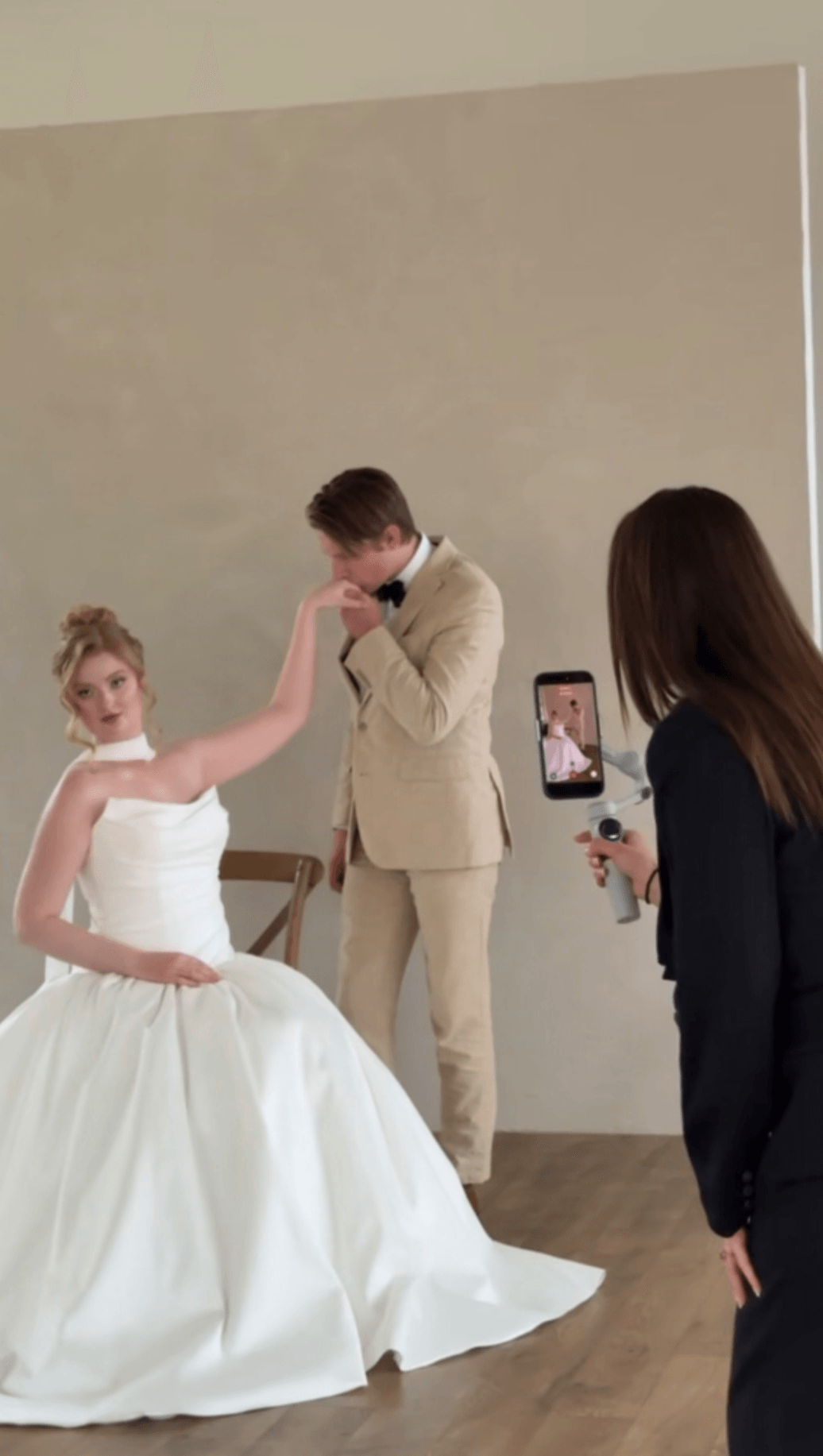 A bride in a white wedding dress and a groom in a beige suit are posing, with a woman taking a photo, in a room with plain walls.