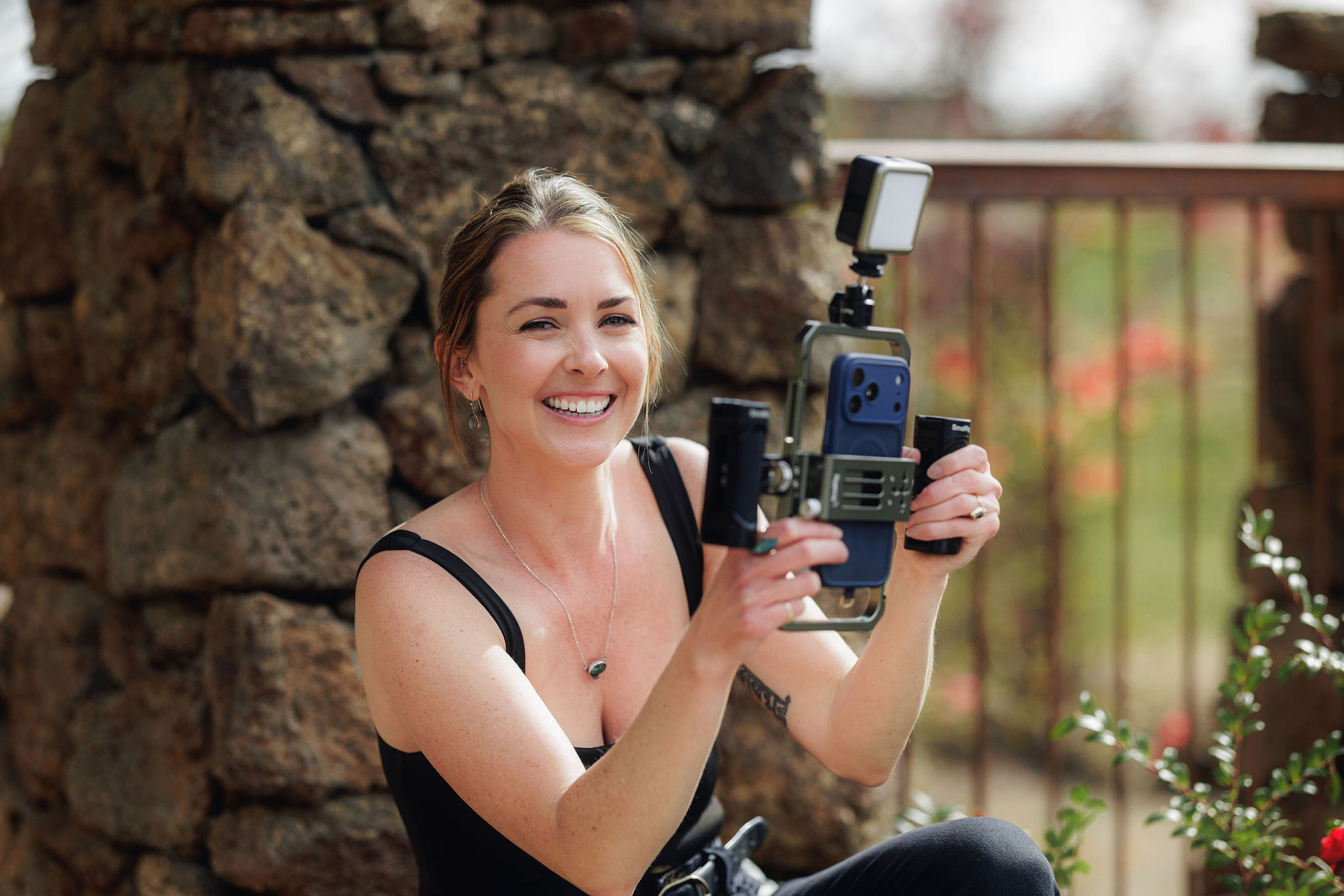 Smiling woman holding a camera with a flash outdoors, stone wall and railing in background.