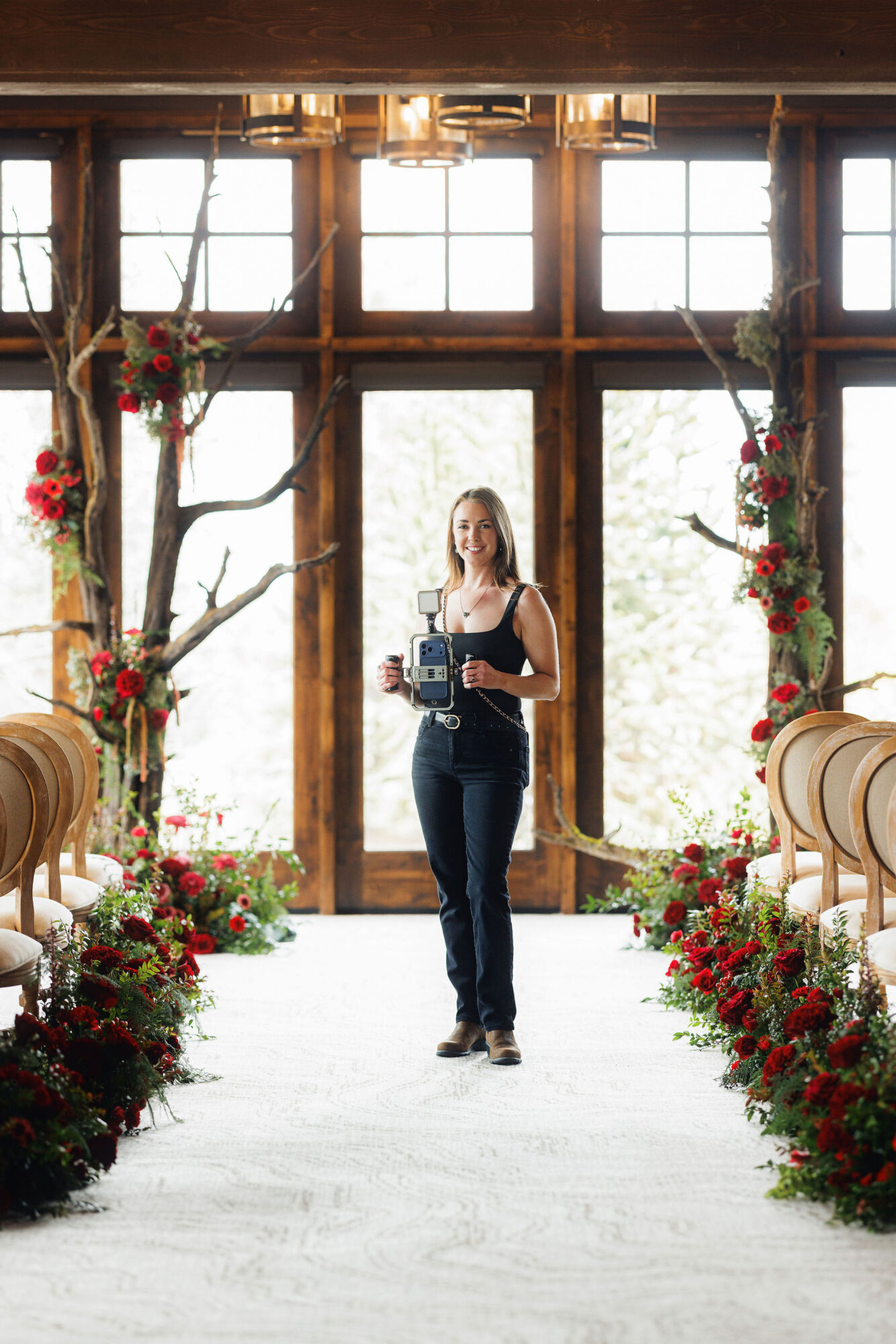 Woman standing in a decorated aisle with large windows behind her, holding a drink, surrounded by floral arrangements and trees.