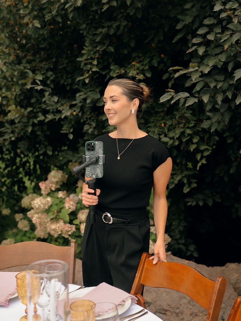 Woman smiling, holding a microphone, standing outdoors with greenery and flowers in background.