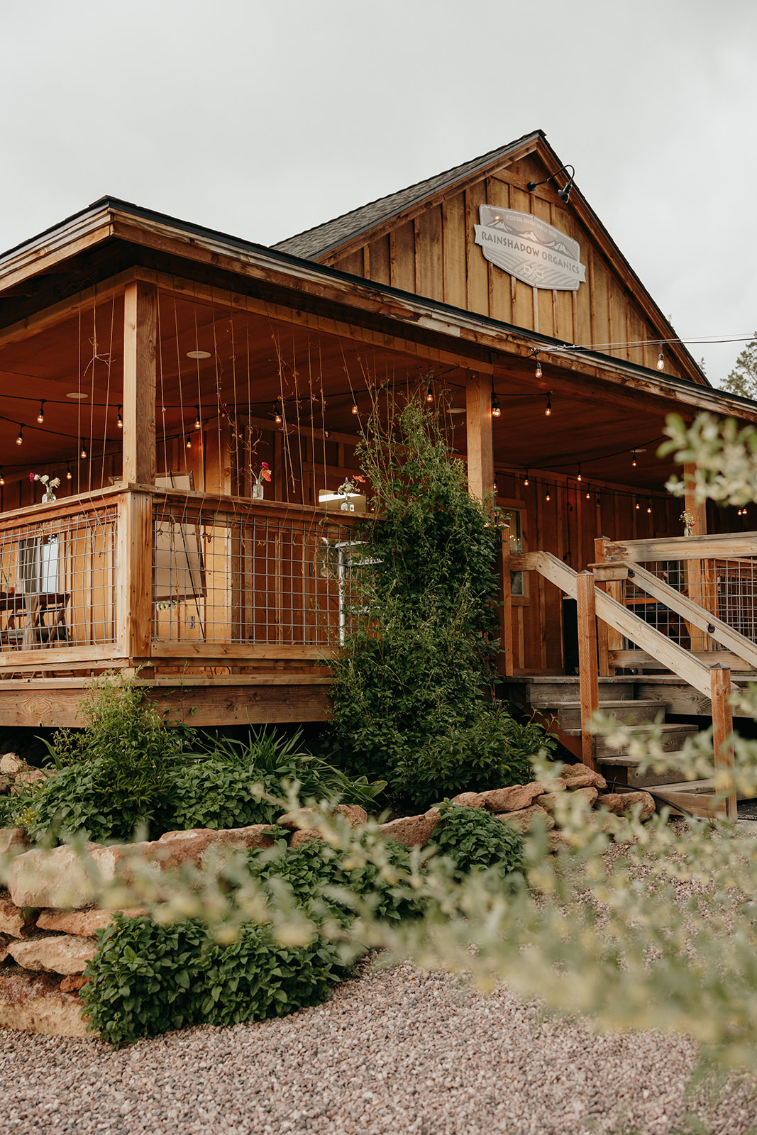 Wooden house with a porch, string lights, and greenery in front, under a cloudy sky.