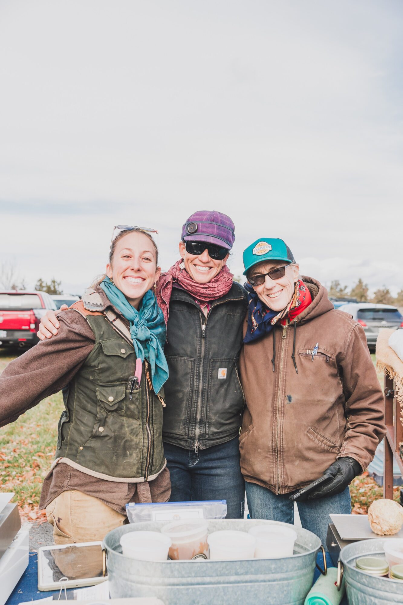Three people smiling outdoors, standing close together with food containers on a table in front of them.