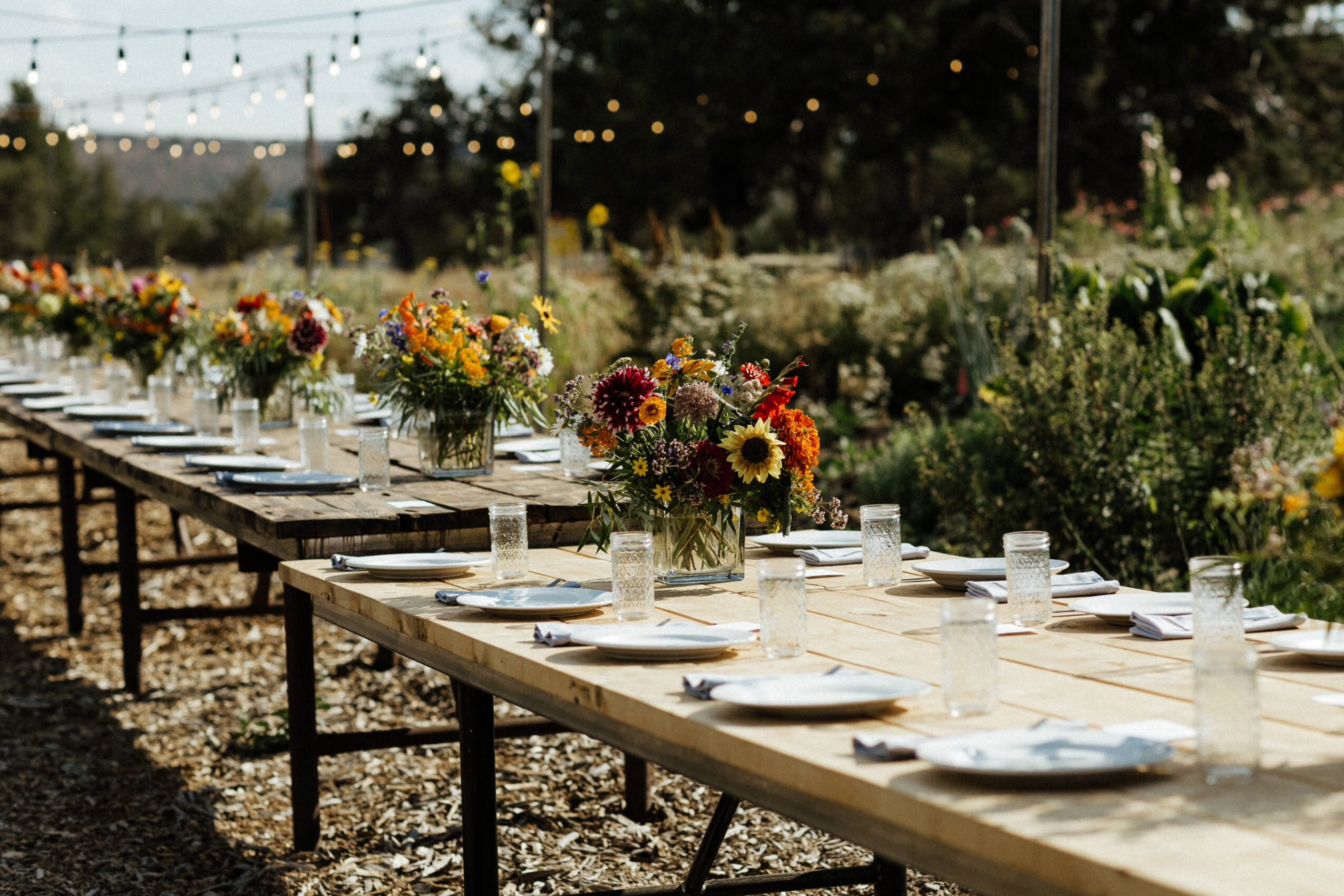 Long outdoor table set with plates, glasses, and colorful flower arrangements, with string lights overhead and greenery in background.