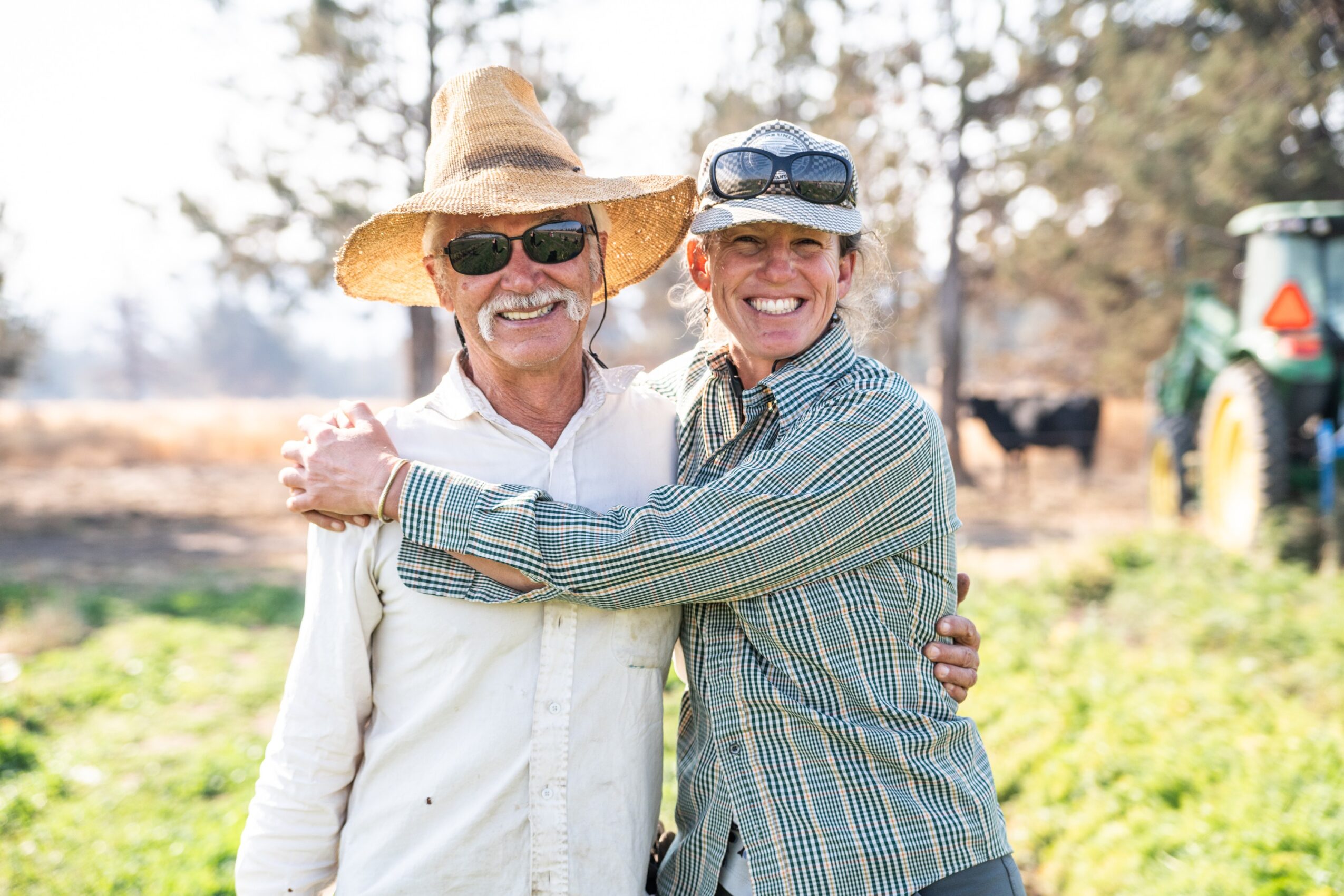 Two people smiling and hugging outdoors, one wearing a large straw hat and sunglasses, the other wearing a cap, in a farm setting