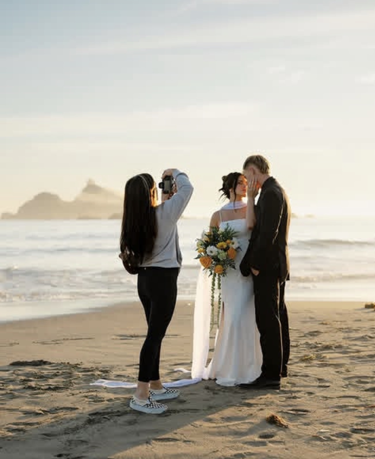 A couple getting married on the beach, with a woman taking a photo, during sunset, holding a bouquet of flowers.