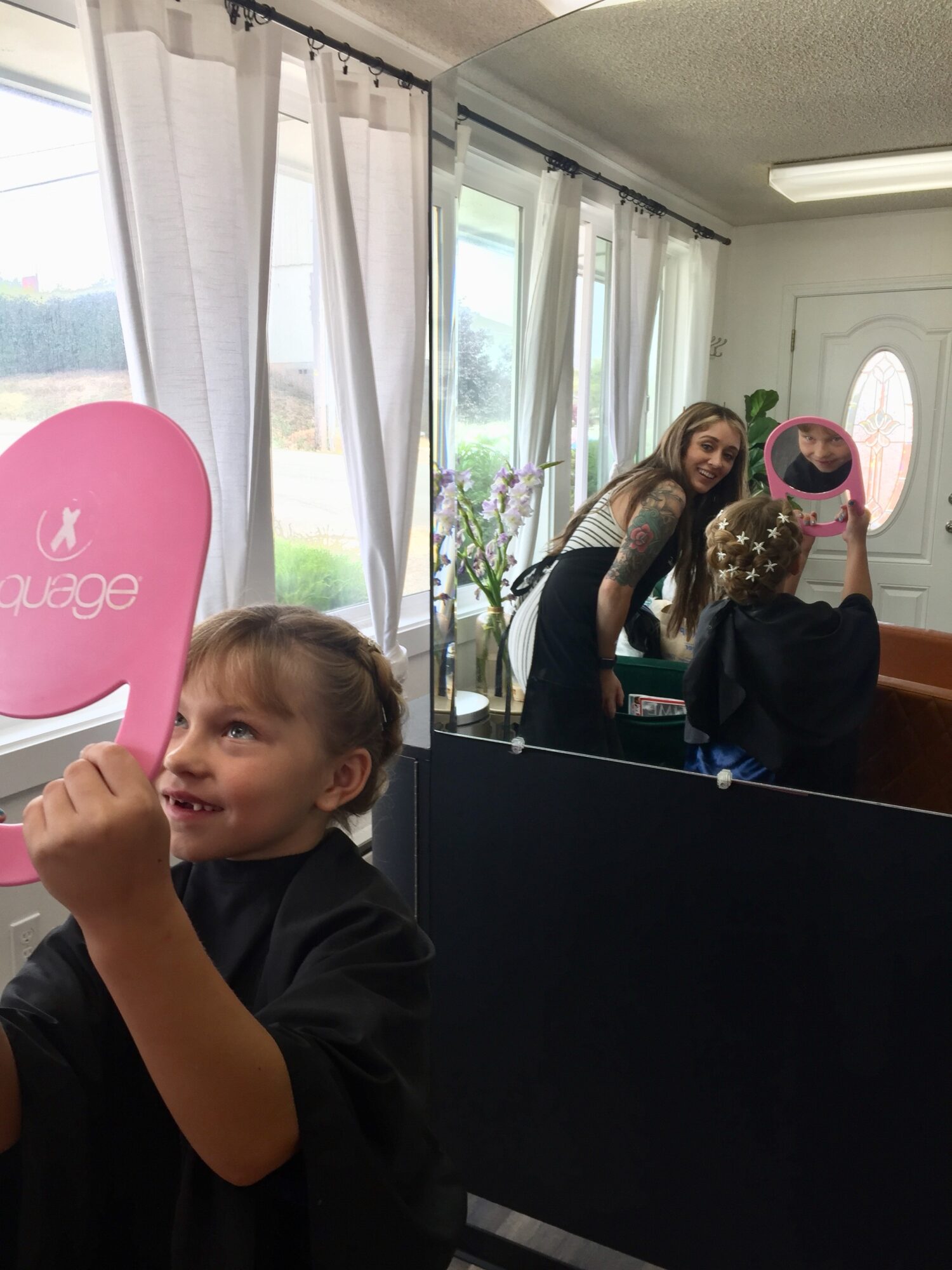 Child smiling and holding a pink balloon, with a woman and another child in a mirror reflection inside a room with large windows and curtains.
