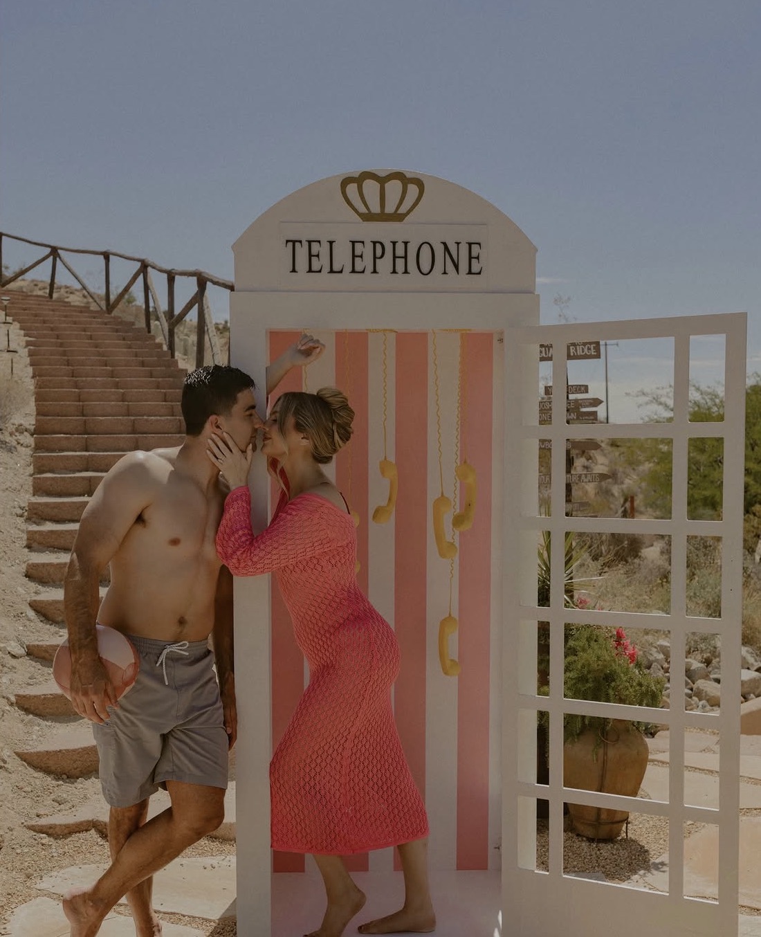 Two people whispering near a decorative telephone booth outdoors, with stairs and plants in the background.