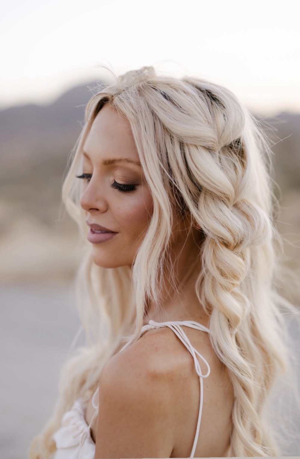 Profile of a woman with long, wavy blonde hair outdoors, looking down, with a blurred natural background.