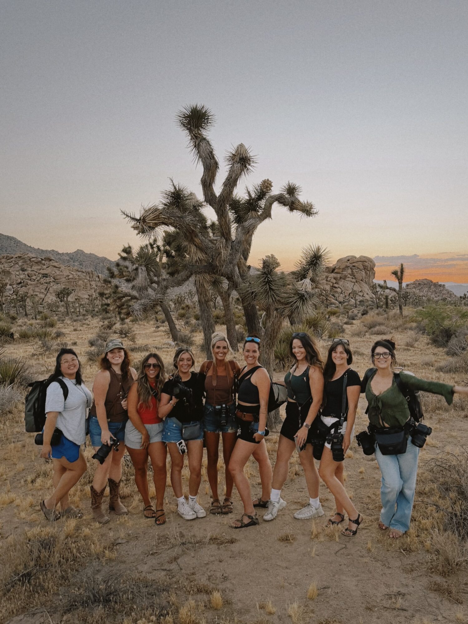 Group of eight people standing outdoors in a desert landscape with a large, twisted tree in the background.