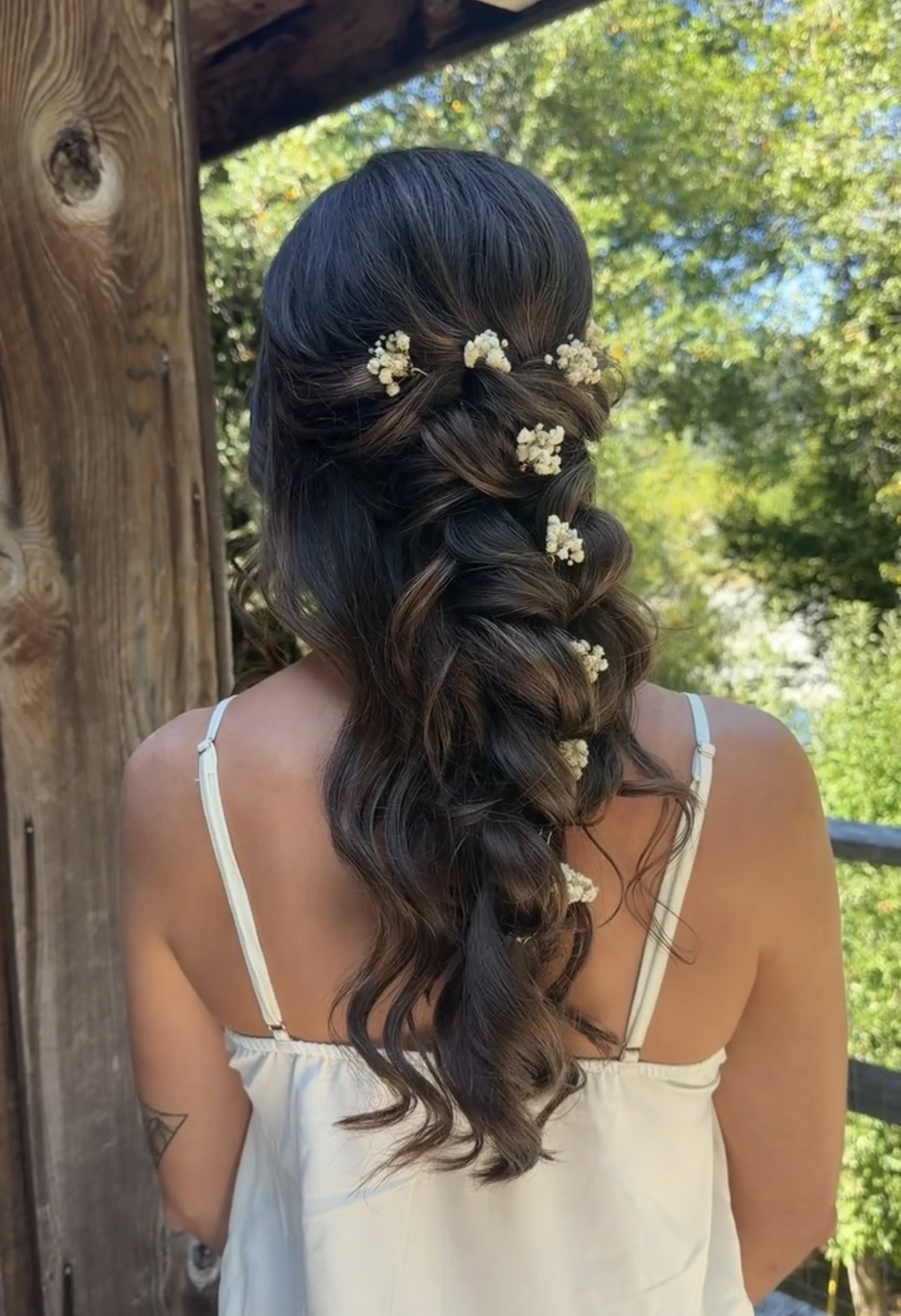 Back view of a woman with long, dark, wavy hair decorated with small white flowers, outdoors with trees and sunlight.