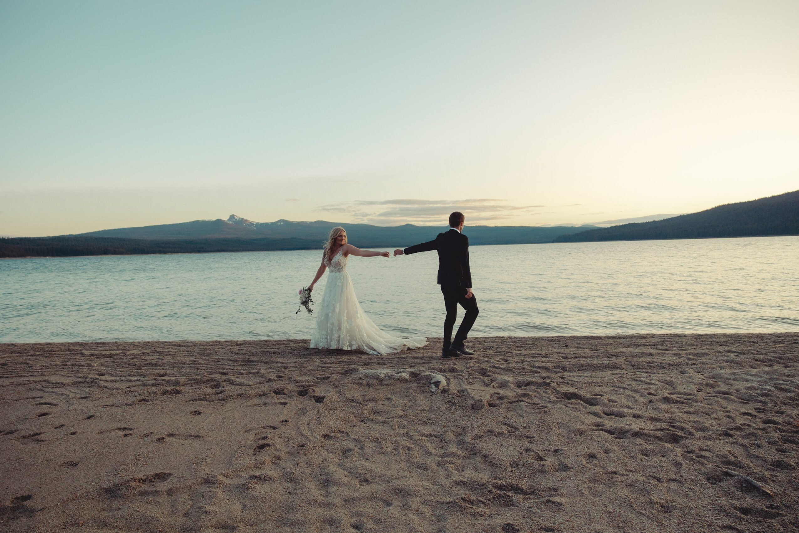 A couple holding hands on a beach at sunset, with water and mountains in the background.