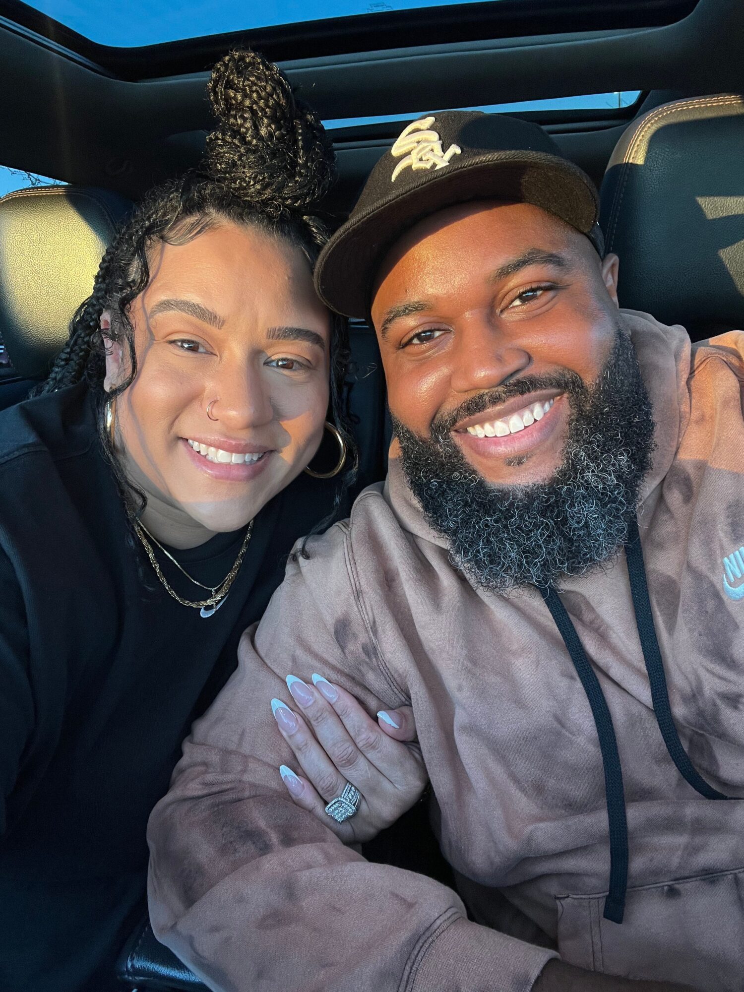 Two smiling people sitting close together, one with a beard and beanie, the other with curly hair, inside a vehicle.