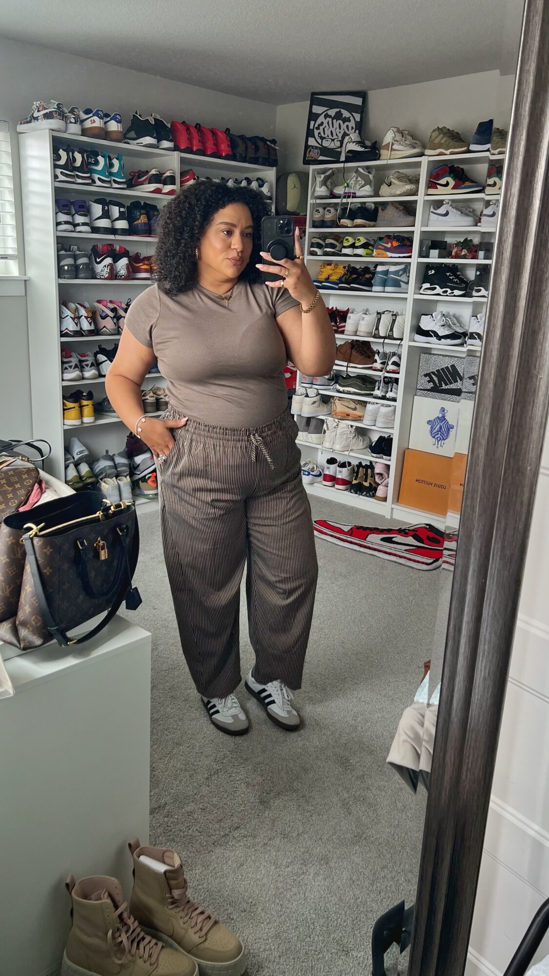Person taking mirror selfie in room with shoe shelves, wearing a beige top and striped pants, holding a phone.