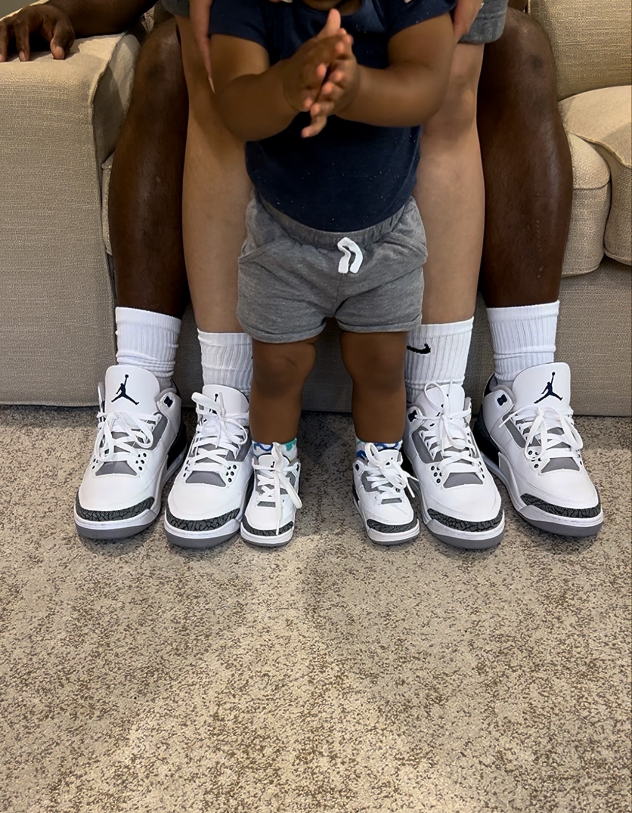 Child standing between two adults, with adults sitting on a couch behind them, wearing sneakers and shorts, on a carpeted floor.
