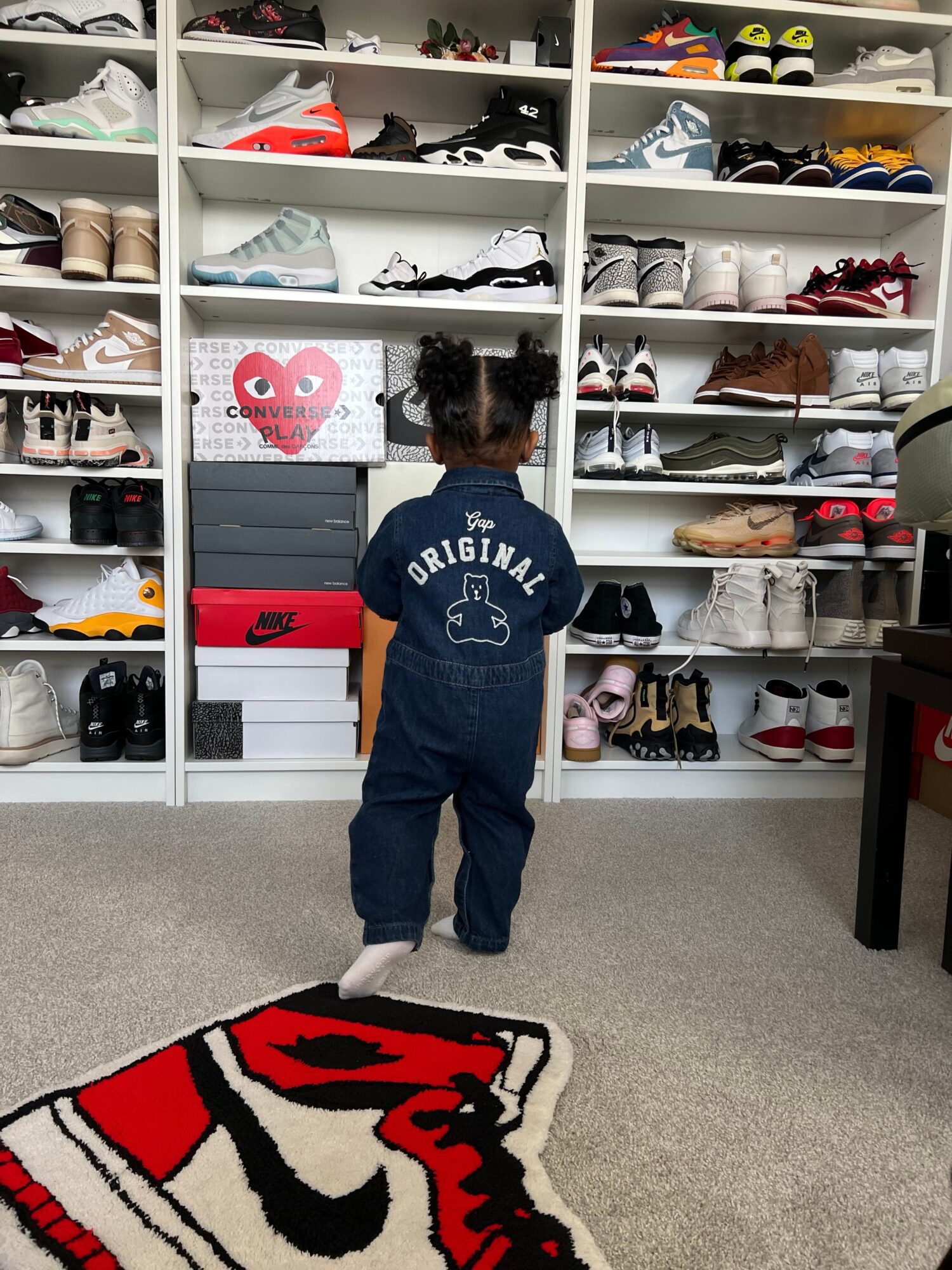 Child with curly hair wearing a navy hoodie and jeans stands in front of a shoe shelf, facing away.