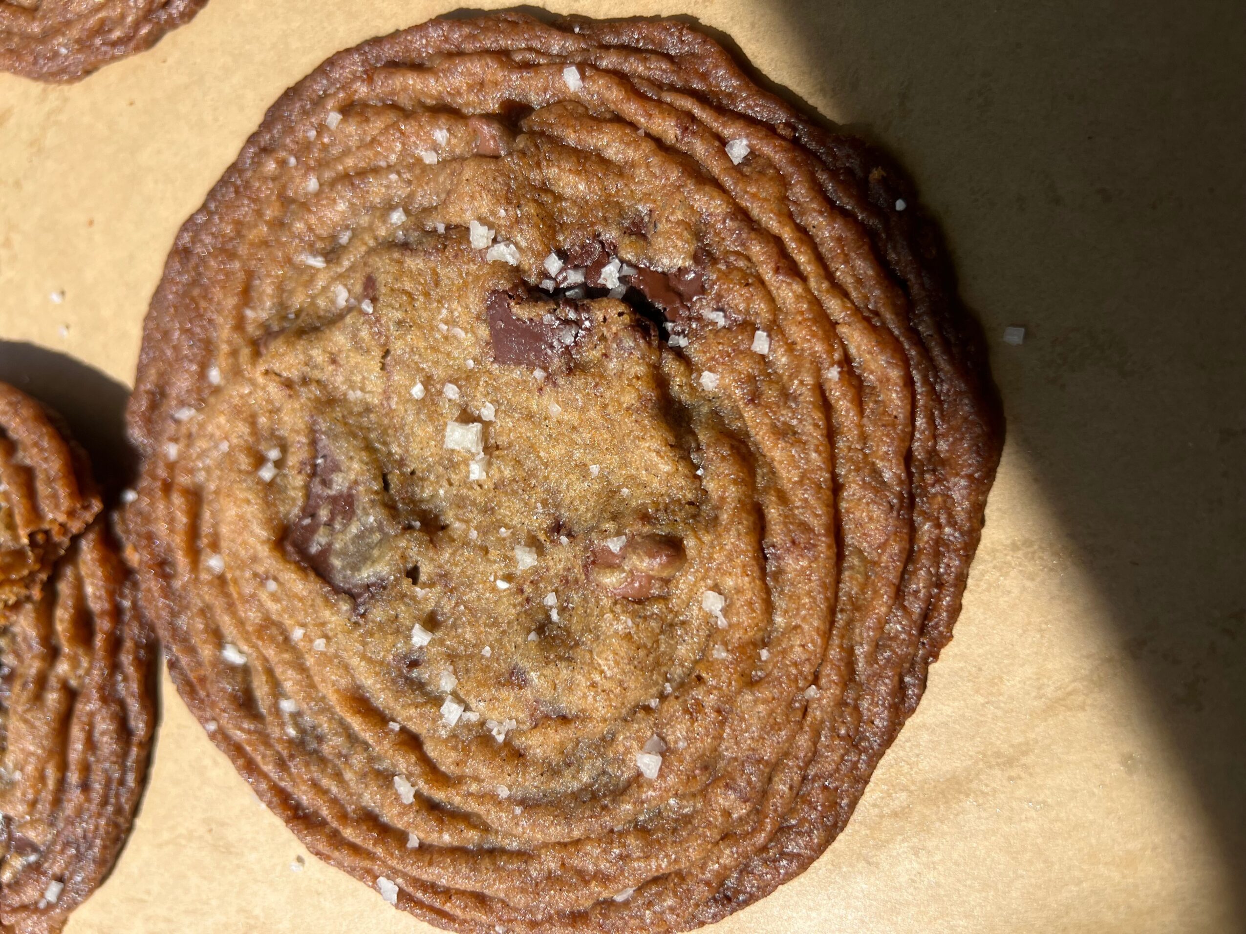 Chocolate chip cookie with salt crystals on top, round and textured, on a light surface with shadow.