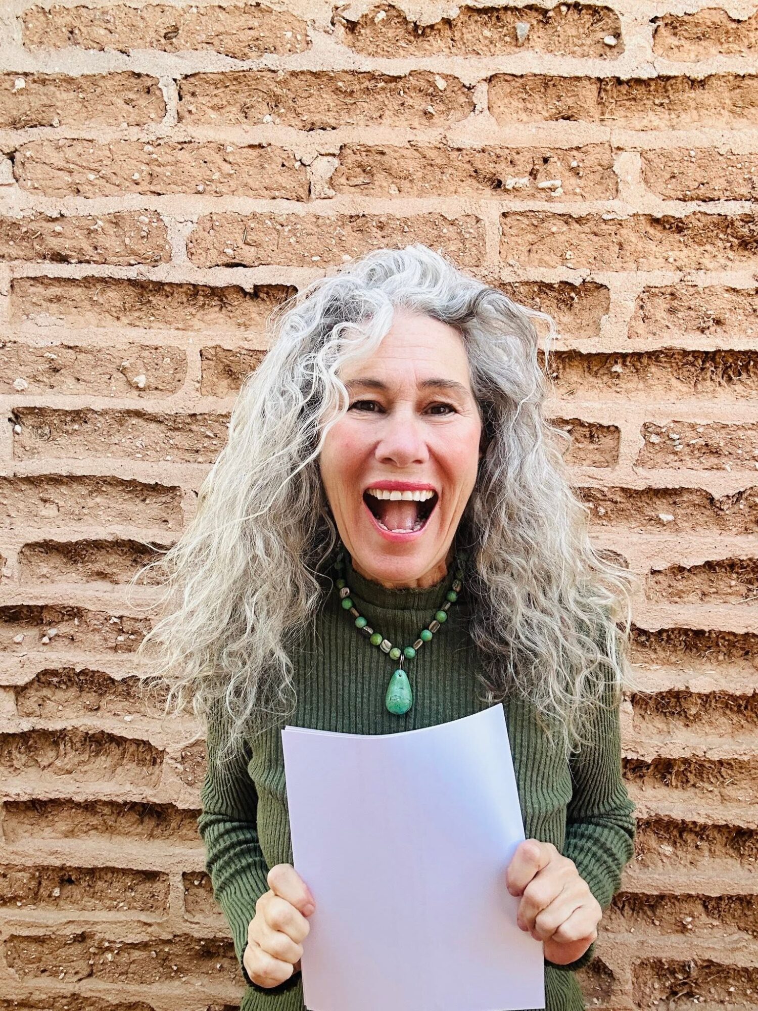 Woman with curly gray hair smiling and holding a blank sheet of paper in front of a brick wall.