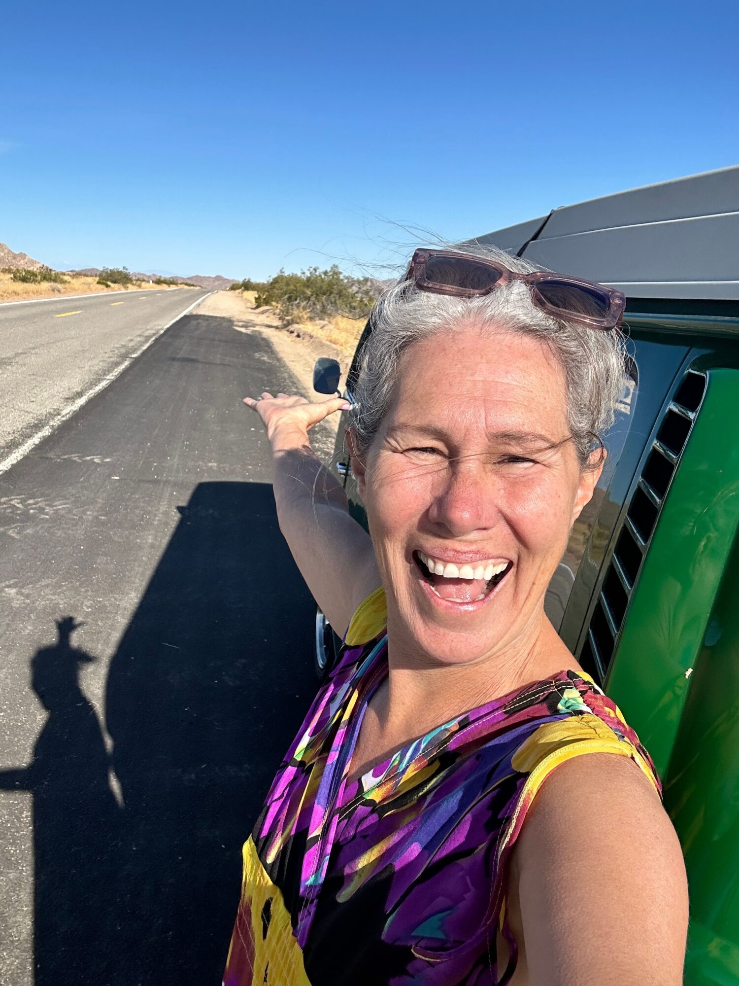 Woman smiling and taking a selfie outdoors on a sunny day, with a parking lot and blue sky in the background.