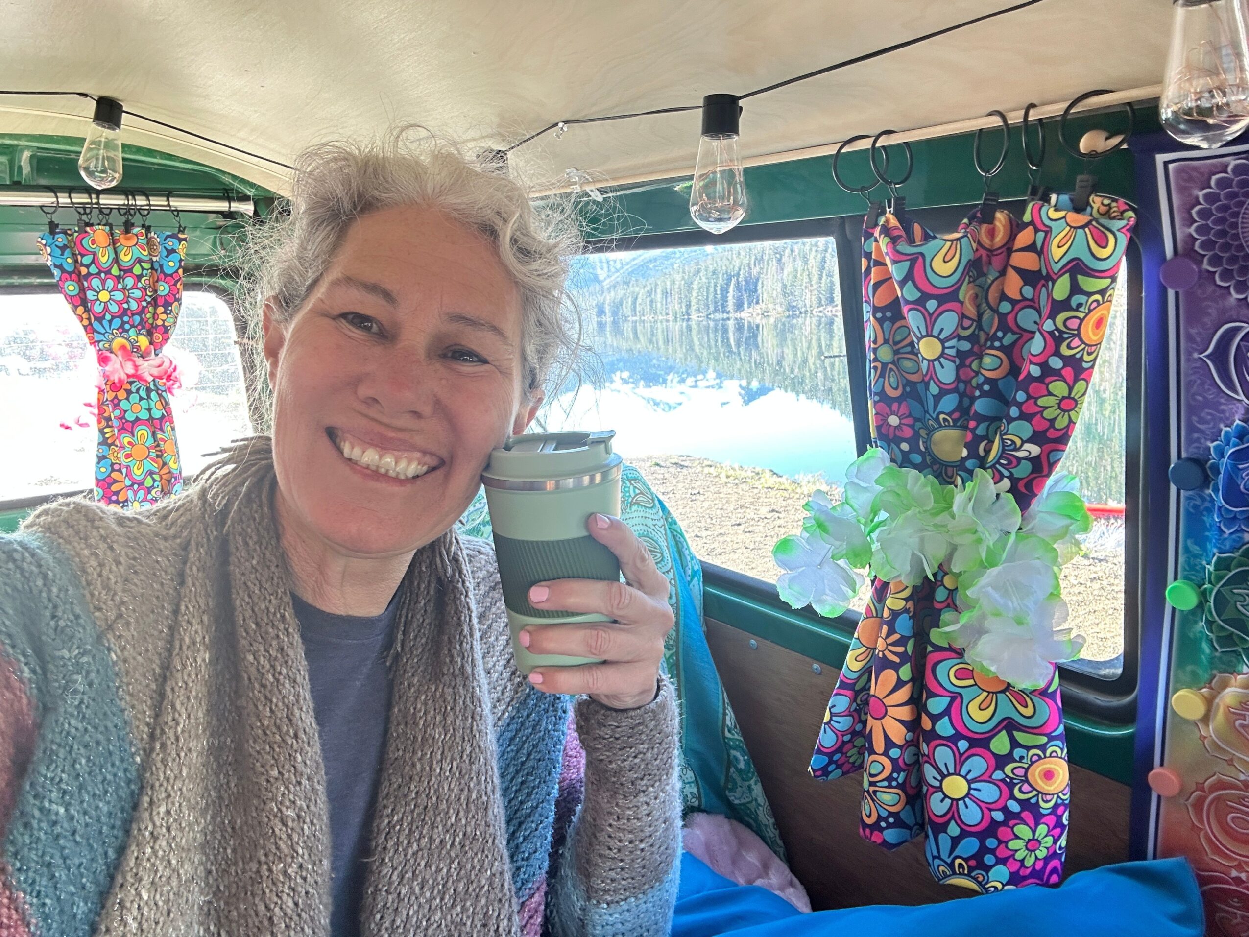 Woman smiling, holding a water bottle, sitting at a table with colorful curtains and a scenic background.