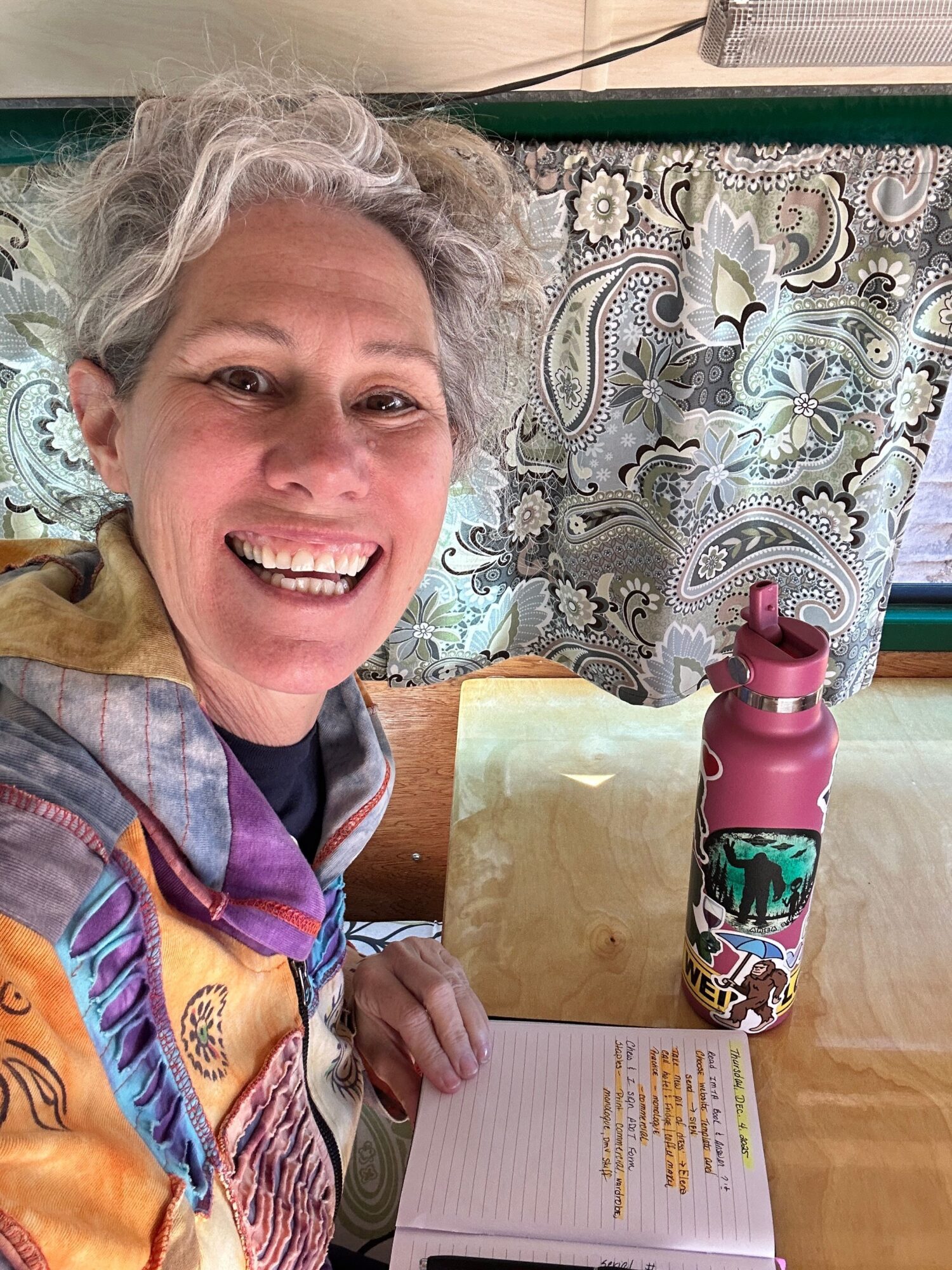 Woman with gray hair smiling, sitting at a table with a notebook and water bottle, in a room with patterned curtains.