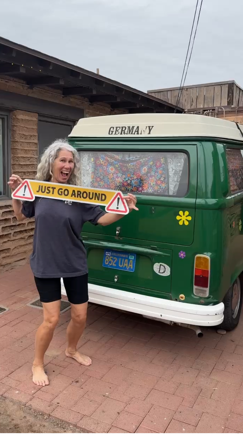 Person holding a yellow sign with red and black text, standing barefoot next to a green van with a yellow flower symbol.