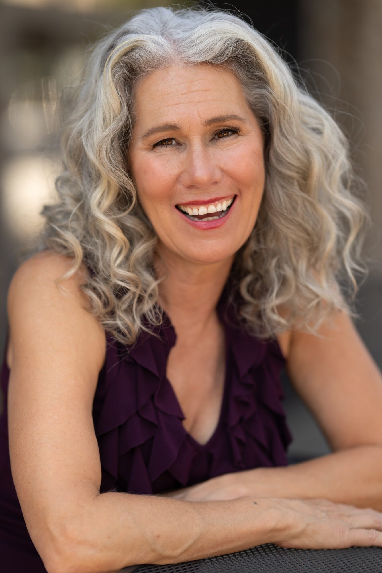 Smiling woman with curly gray hair wearing a purple top, sitting outdoors with blurred background.