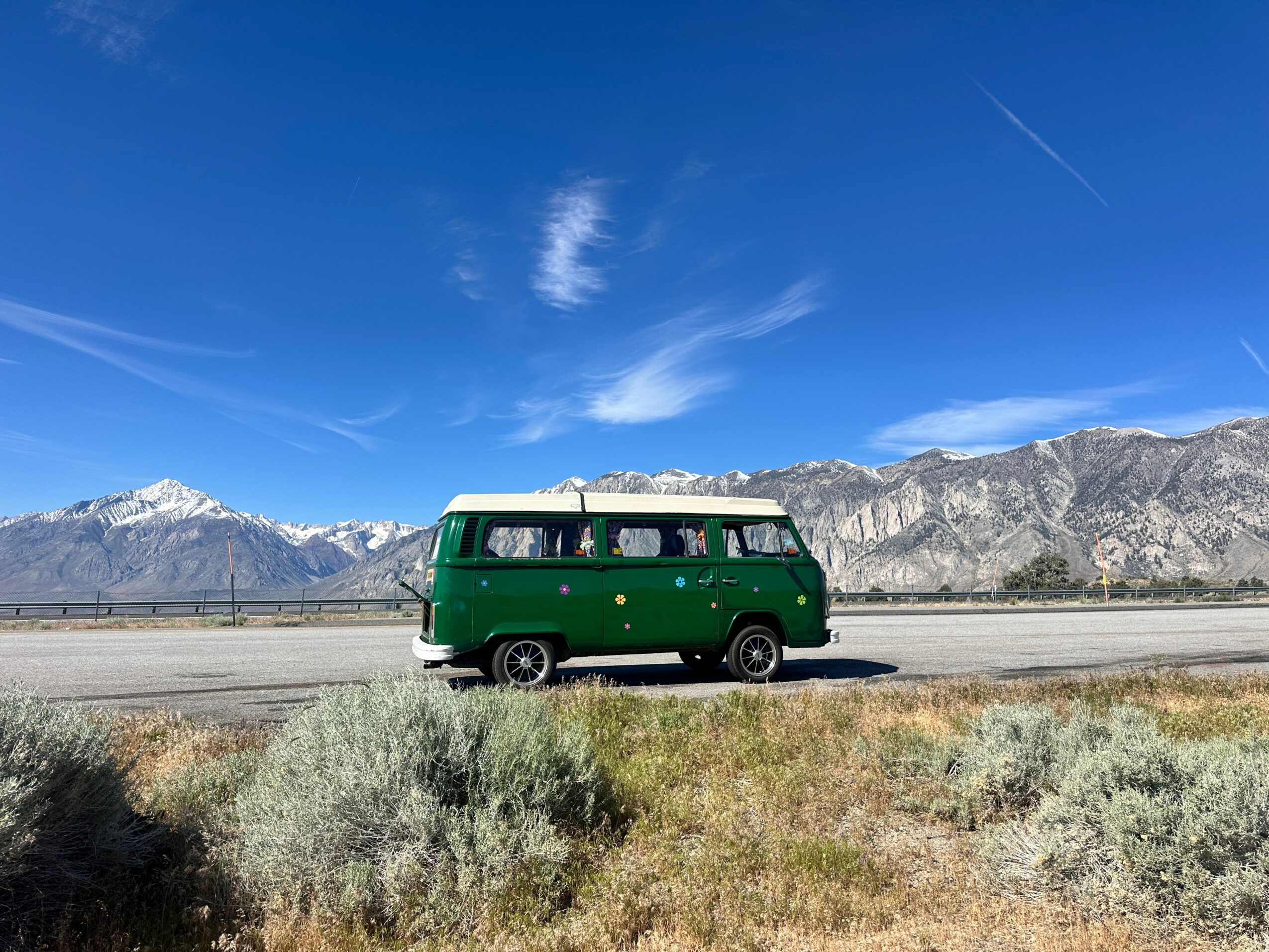 Green van driving on a road with mountains and blue sky in the background.