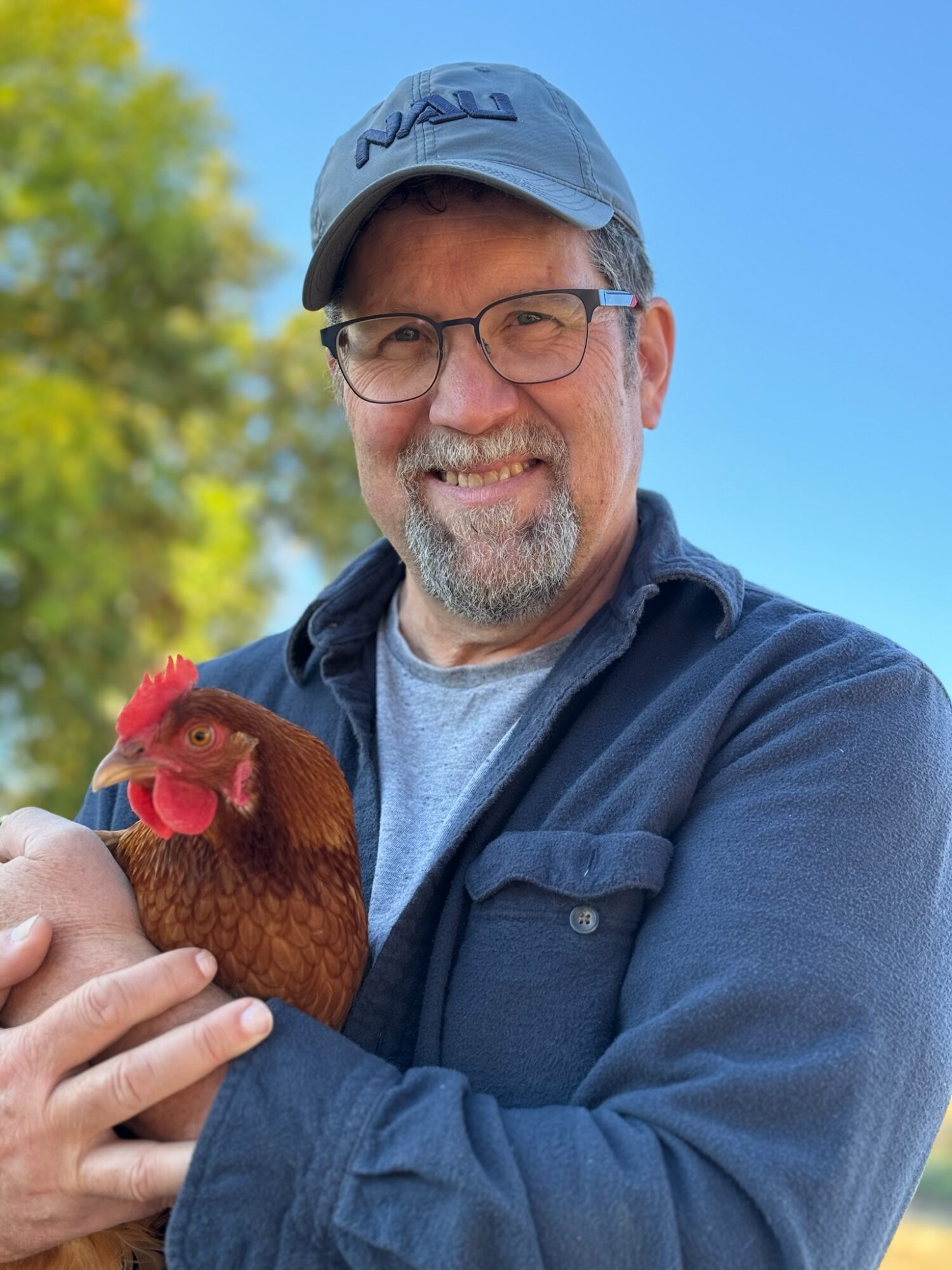 Man holding a chicken outdoors, smiling, wearing glasses, a cap, and a jacket, with trees and blue sky in background.