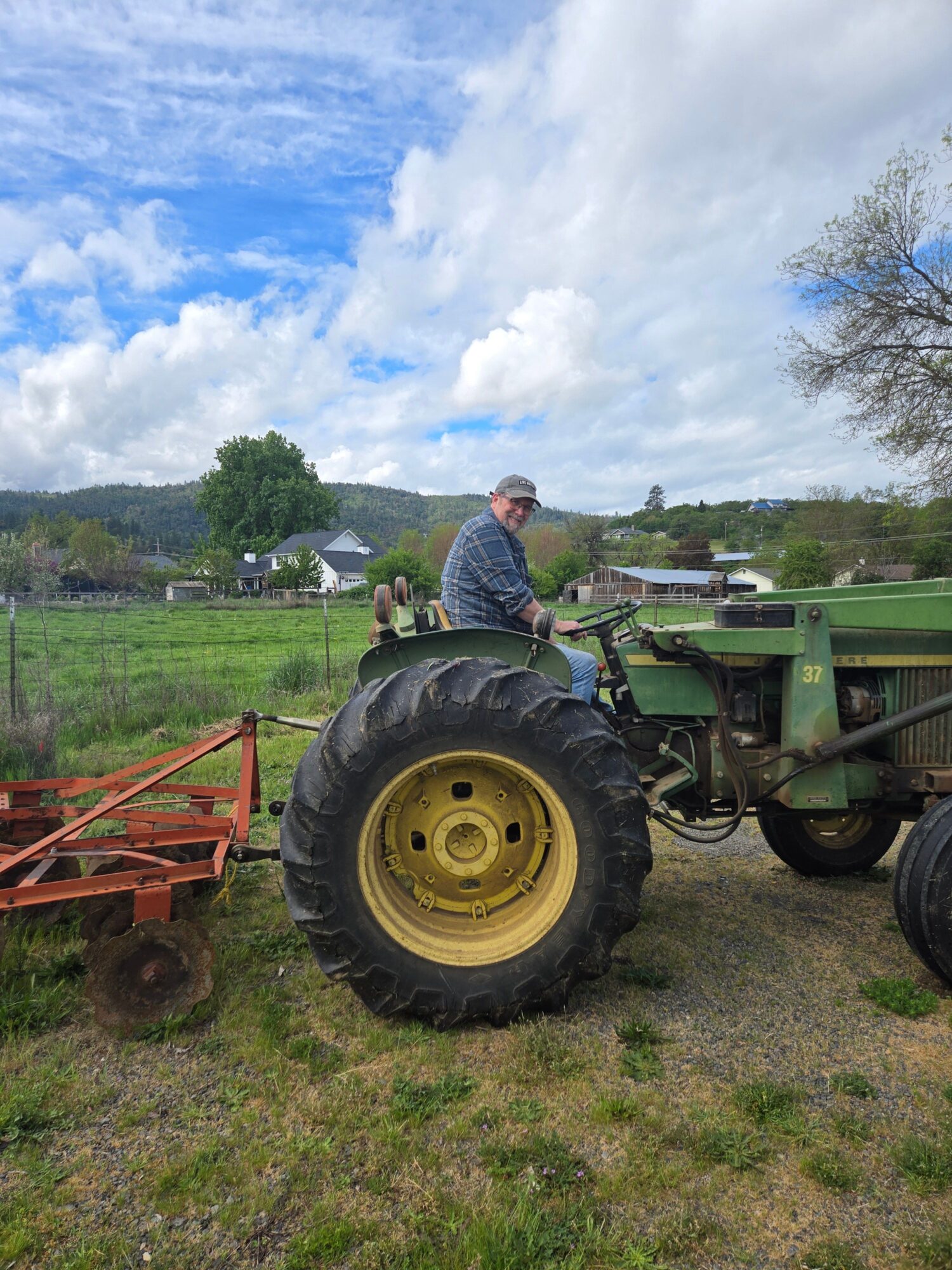 Person sitting on a green tractor in a grassy field with trees and cloudy sky in the background.