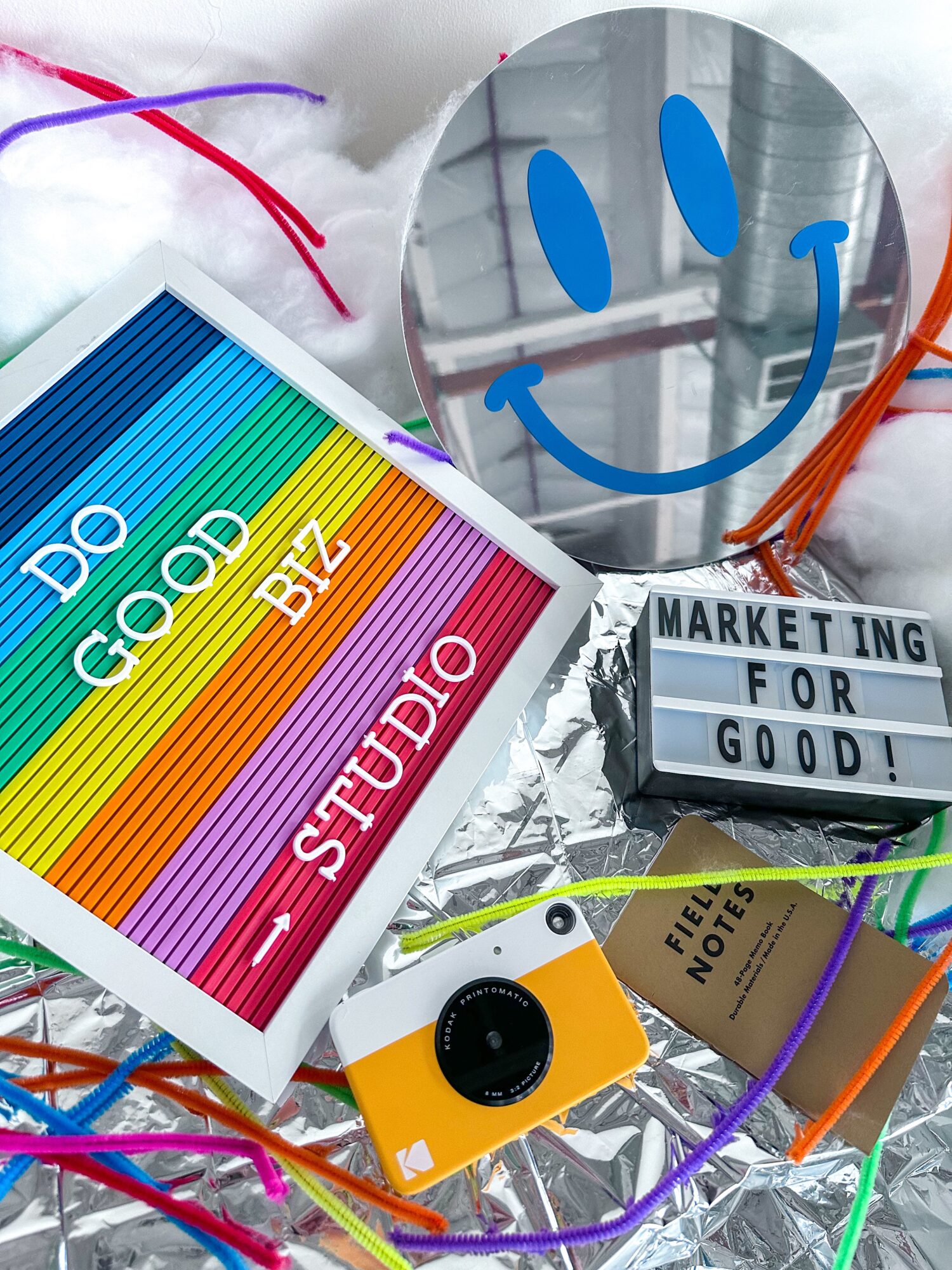 A mirror with a smiley face, colorful sign reading 'Do Good' and 'Studio,' a camera, and a sign saying 'Marketing for Good,' on a table.